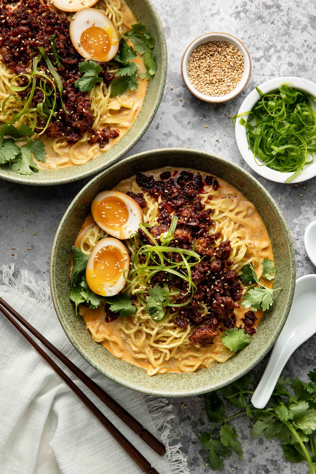 Two bowls of ramen topped with soft-boiled eggs, chili oil, fresh cilantro, and green onions. Sliced green onions and sesame seeds are in small bowls nearby. Chopsticks and a white napkin are placed beside the bowls.