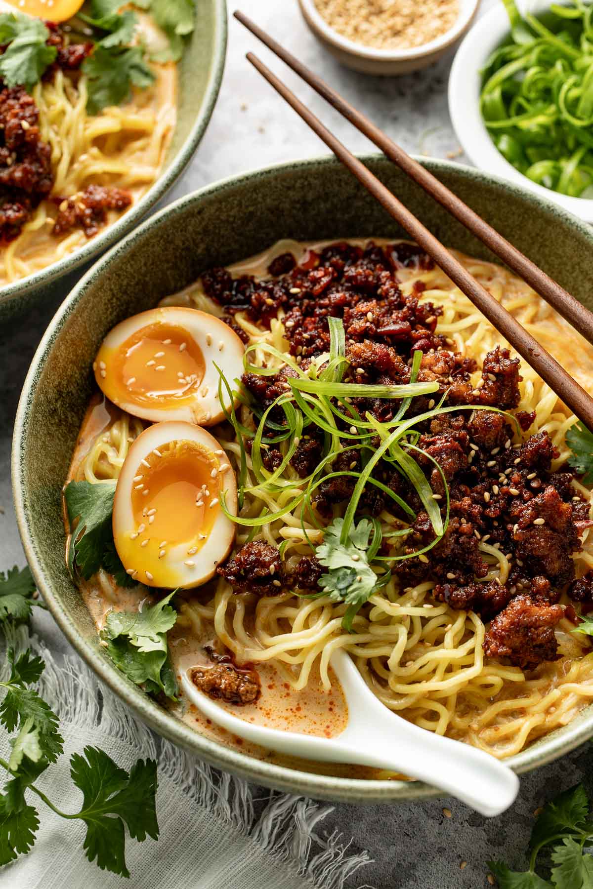 A bowl of ramen topped with soft-boiled eggs, minced meat, green onions, cilantro, and sesame seeds, with chopsticks and a ceramic spoon on the side. Garnishes and extra toppings are visible in the background.