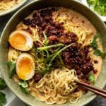 A bowl of ramen with noodles, halved soft-boiled egg, spicy minced meat, fresh greens, and broth, garnished with green onions. Chopsticks and small bowls of sesame seeds and scallions are nearby.