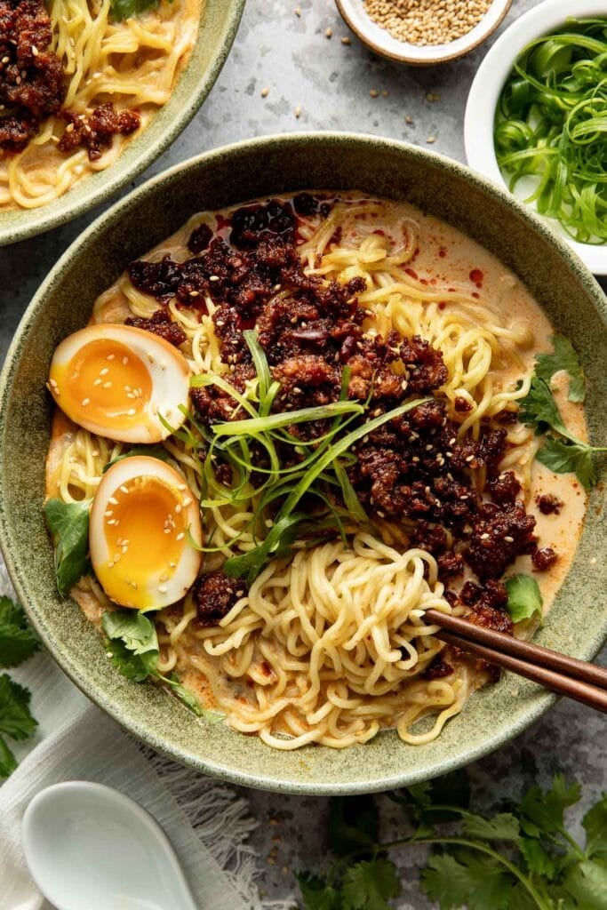 A bowl of ramen with noodles, halved soft-boiled egg, spicy minced meat, fresh greens, and broth, garnished with green onions. Chopsticks and small bowls of sesame seeds and scallions are nearby.