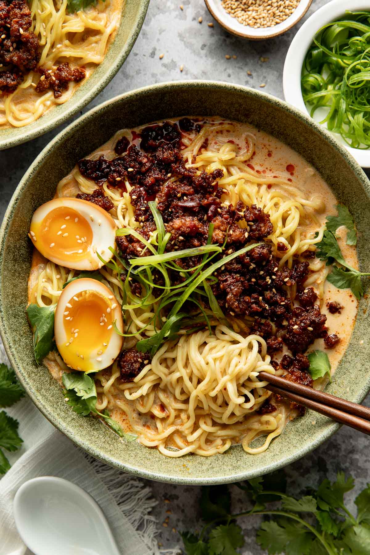 A bowl of ramen with noodles, halved soft-boiled egg, spicy minced meat, fresh greens, and broth, garnished with green onions. Chopsticks and small bowls of sesame seeds and scallions are nearby.