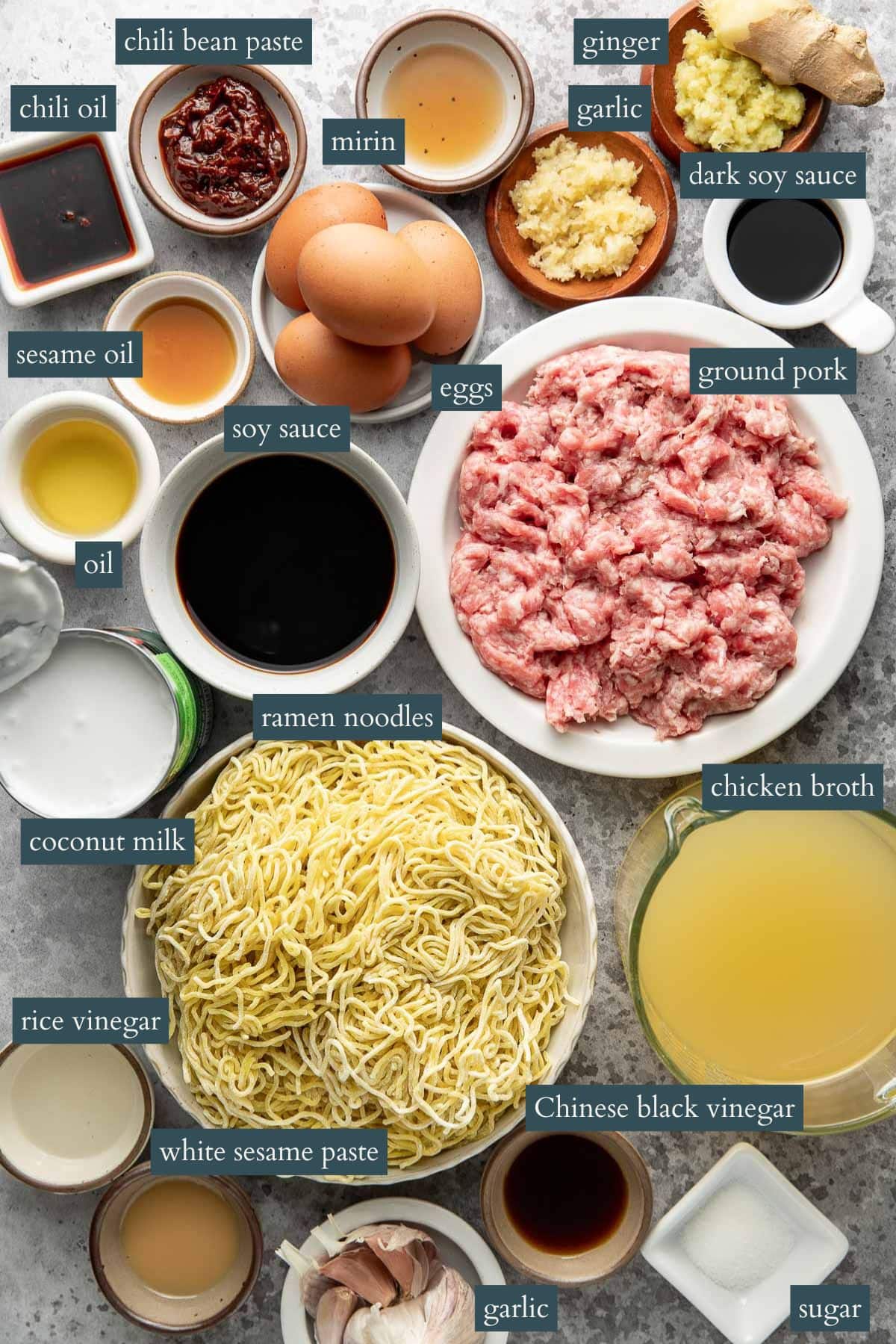 Various labeled ingredients for making ramen, including ground pork, ramen noodles, eggs, soy sauce, chicken broth, coconut milk, garlic, ginger, chili oil, and condiments, arranged on a countertop.