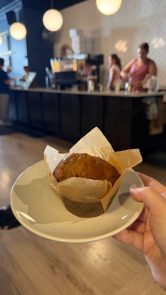 A hand holds a plate with a muffin in a paper liner, while a coffee shop counter and baristas are visible in the blurred background.