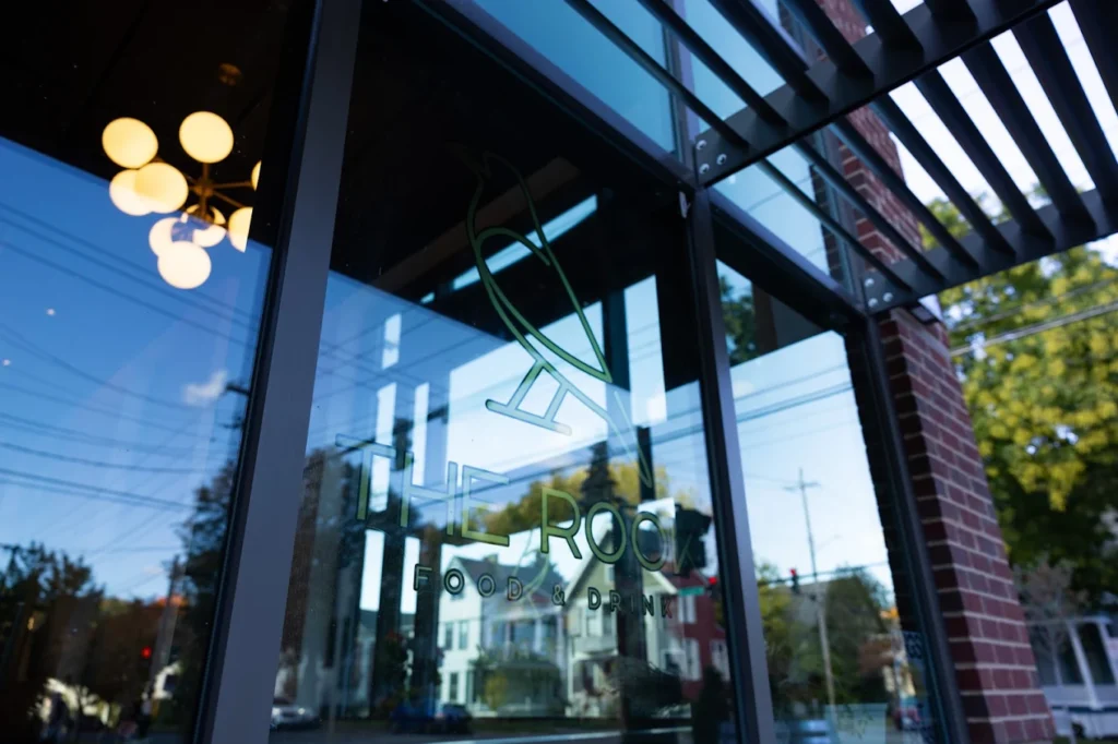 A glass storefront with the logo of The Roost Food & Drink, reflecting trees, buildings, and power lines outside. Modern light fixtures and metal awnings are visible, creating a welcoming atmosphere in this Finger Lakes destination.
