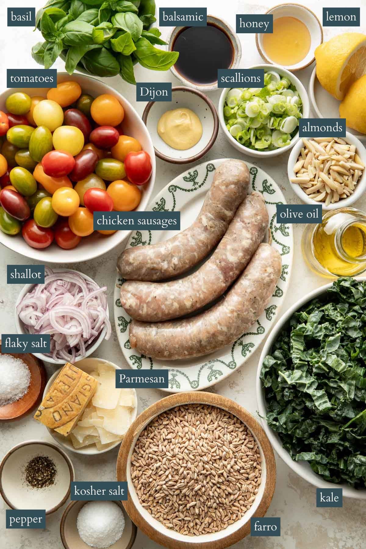 An overhead view of ingredients labeled: basil, balsamic, honey, lemon, tomatoes, Dijon, scallions, almonds, chicken sausages, olive oil, shallot, flaky salt, kosher salt, pepper, Parmesan, farro, and kale.