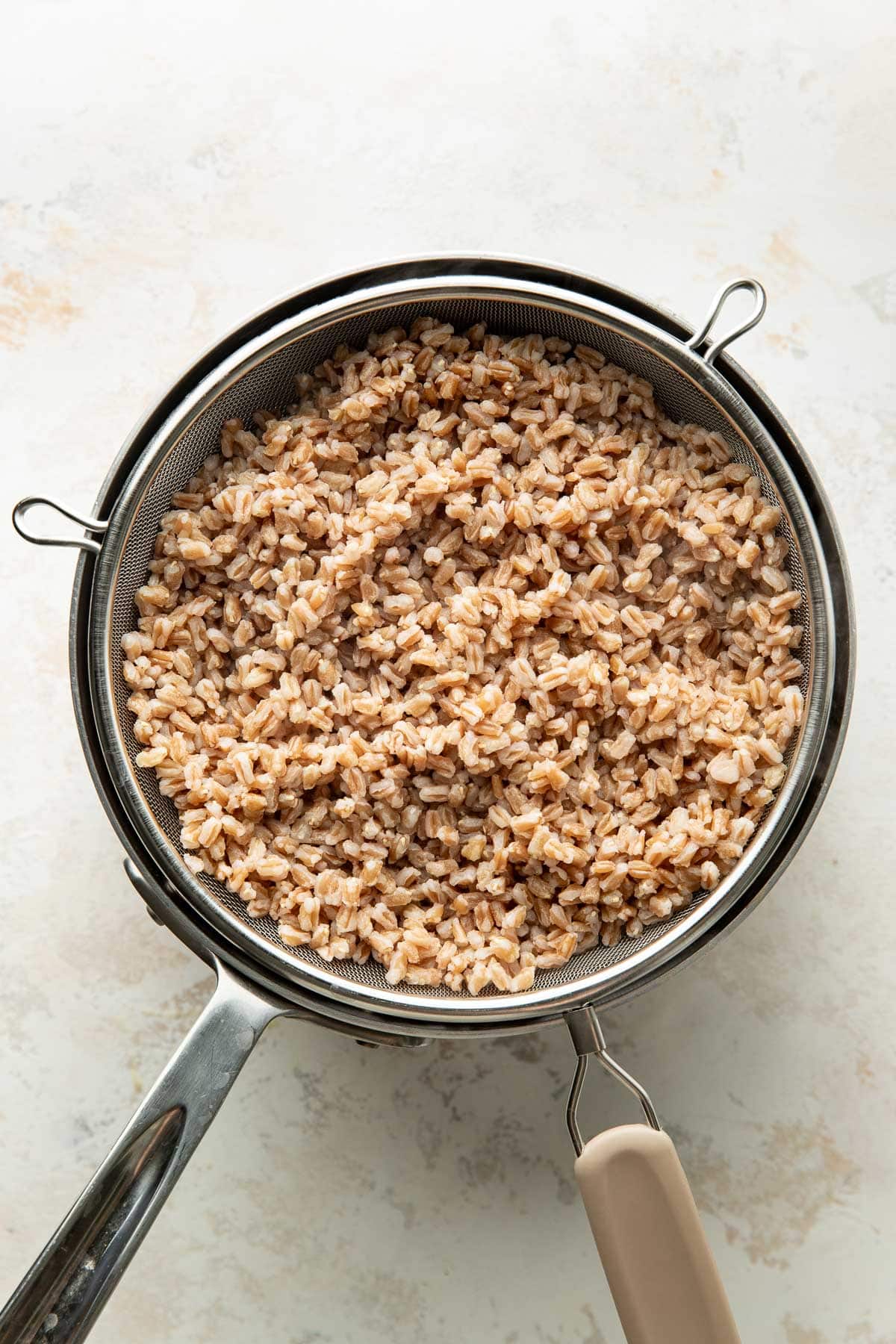 Cooked farro draining in a metal fine-mesh strainer placed over a saucepan, on a light-colored surface. The farro grains appear light brown and slightly glossy.