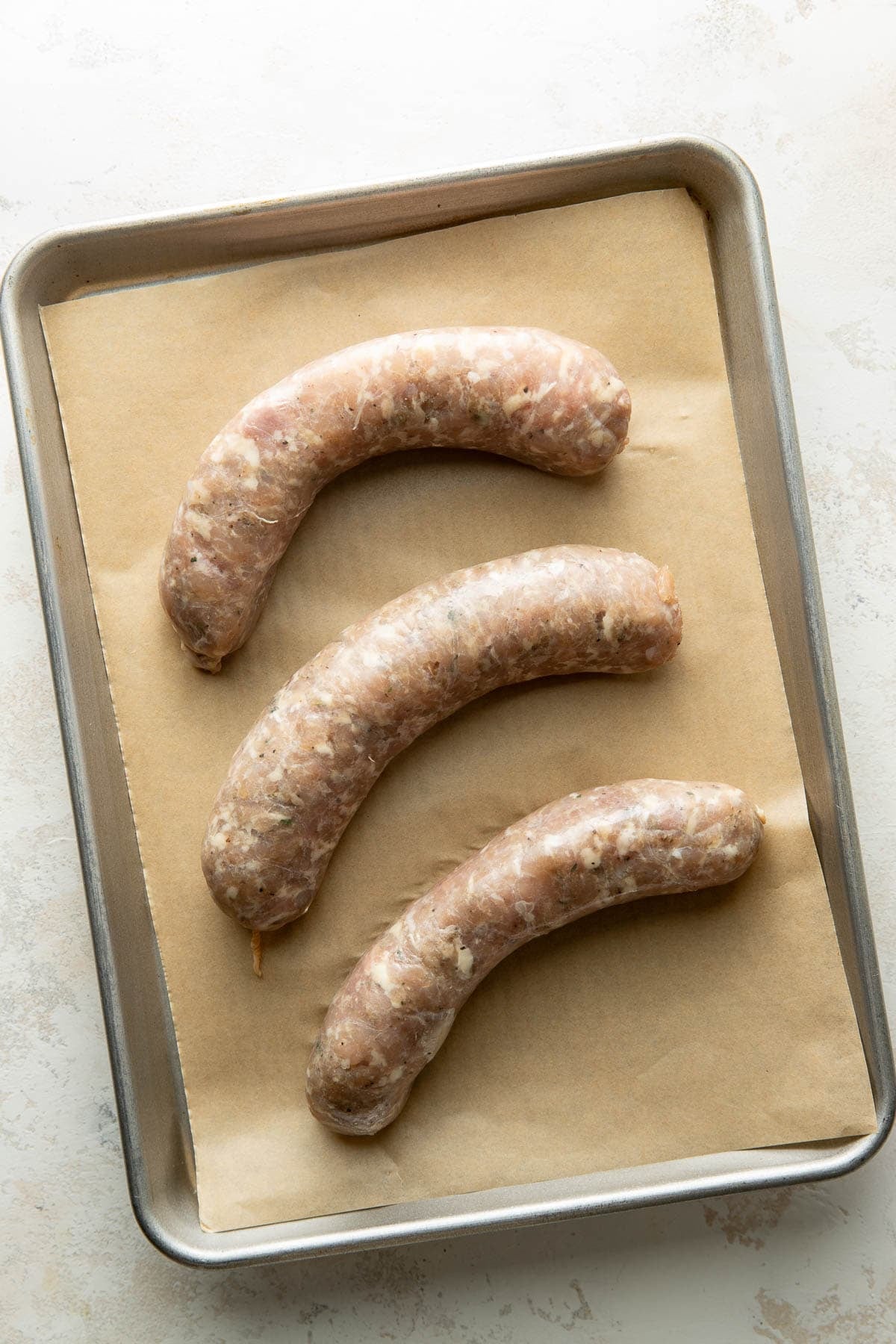 Three raw sausages arranged in a row on a parchment-lined baking sheet, ready for cooking.