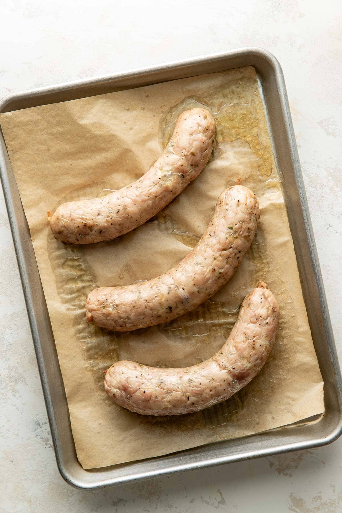 Three raw sausages placed on a parchment-lined baking sheet, arranged with space between them, ready for cooking.