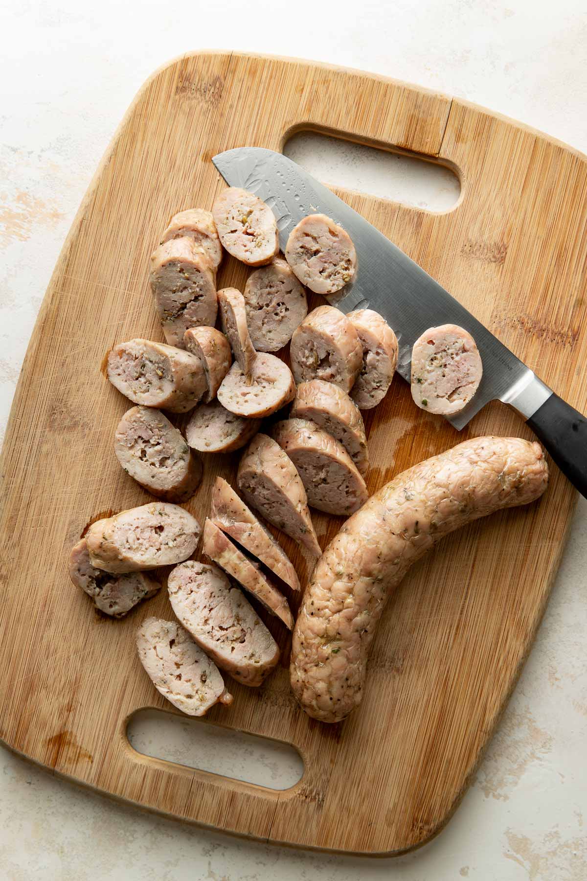 A sliced sausage and a whole sausage are on a wooden cutting board next to a large knife. The sausage pieces are arranged in a pile. The scene is well-lit and viewed from above.