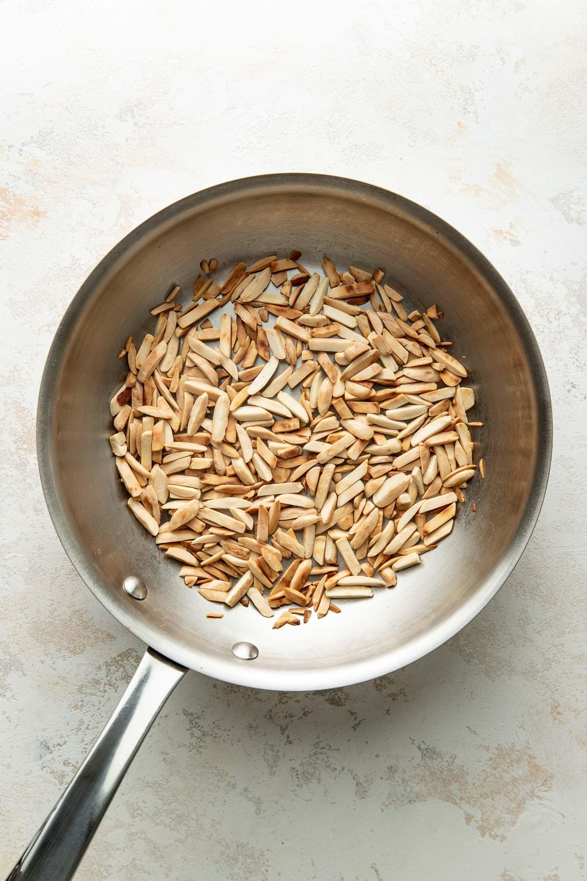 Slivered almonds are being toasted in a stainless steel pan on a light-colored surface.