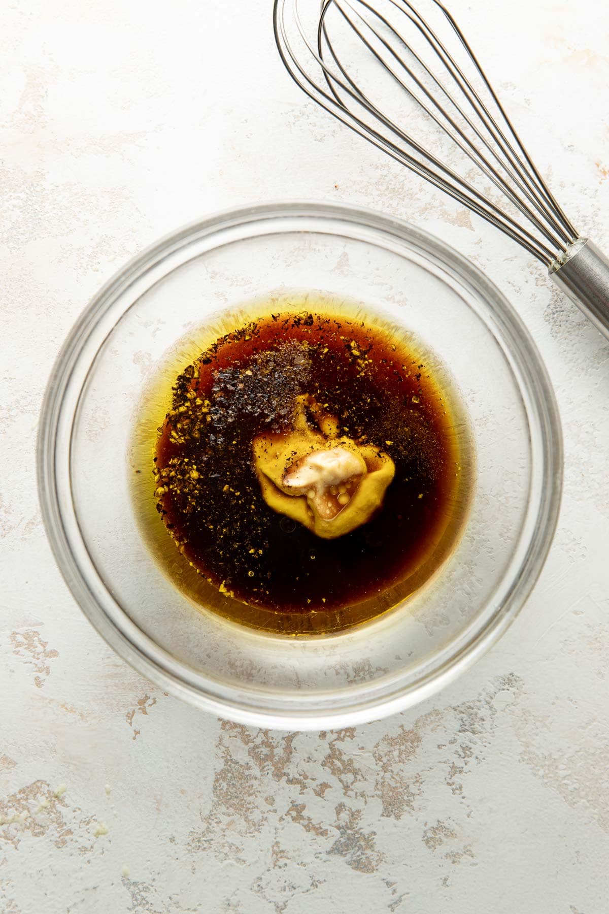 A glass bowl with mustard, soy sauce, pepper, and other seasonings, next to a metal whisk on a light-colored surface.