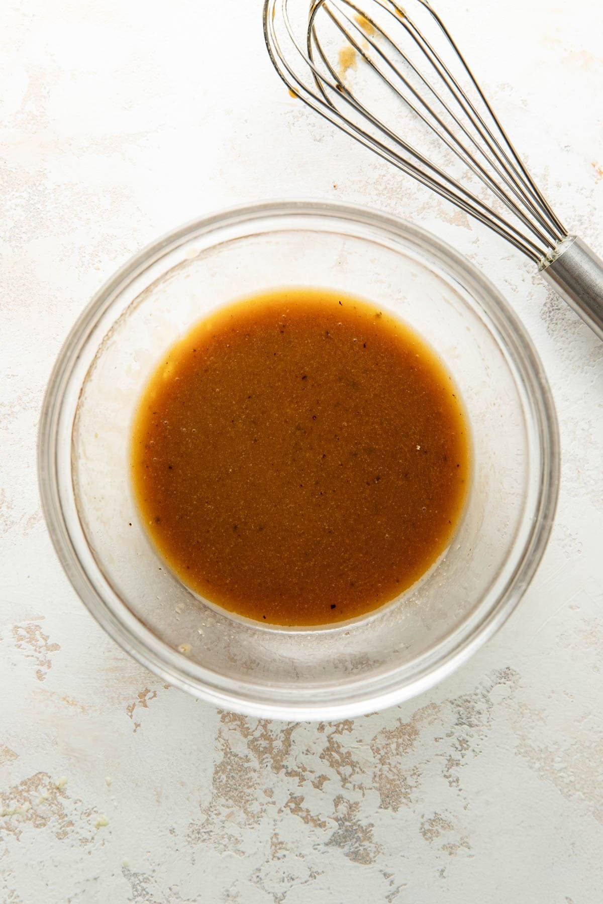 A glass bowl of brown vinaigrette sits on a light surface, with a metal whisk placed nearby.