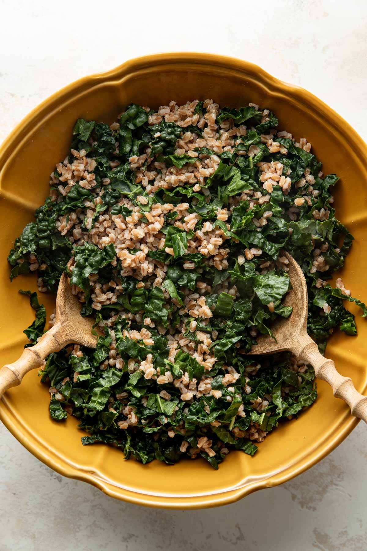 A yellow bowl filled with a mixture of chopped kale and cooked brown rice, with two wooden serving utensils resting inside.