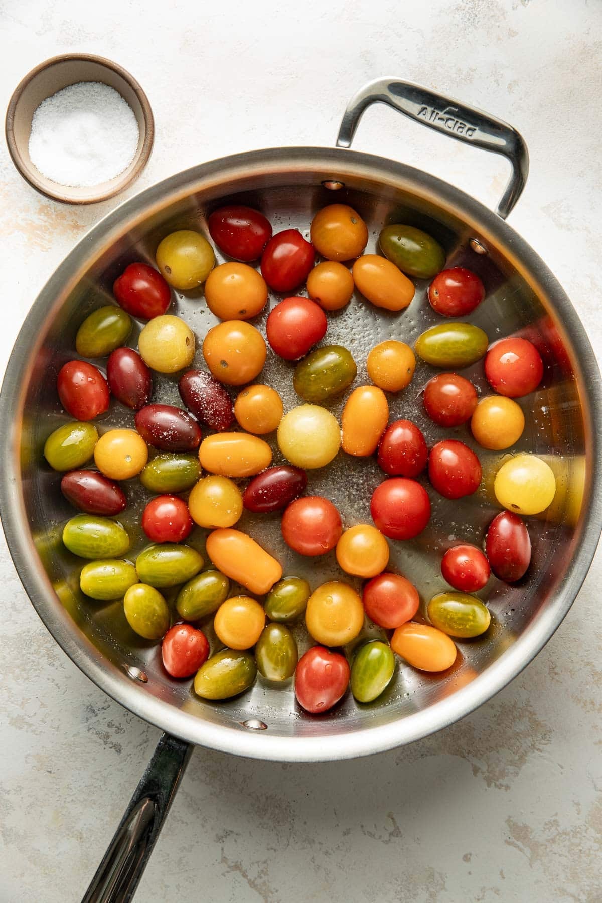A stainless steel pan filled with various colorful cherry tomatoes sits on a light surface. A small bowl of coarse salt is nearby.