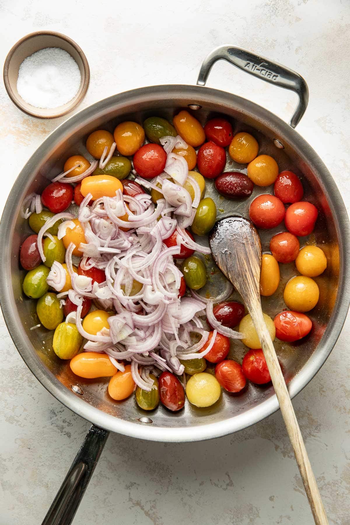 A skillet filled with colorful cherry tomatoes and sliced shallots, being stirred with a wooden spoon. A small bowl of salt sits nearby on a light countertop.