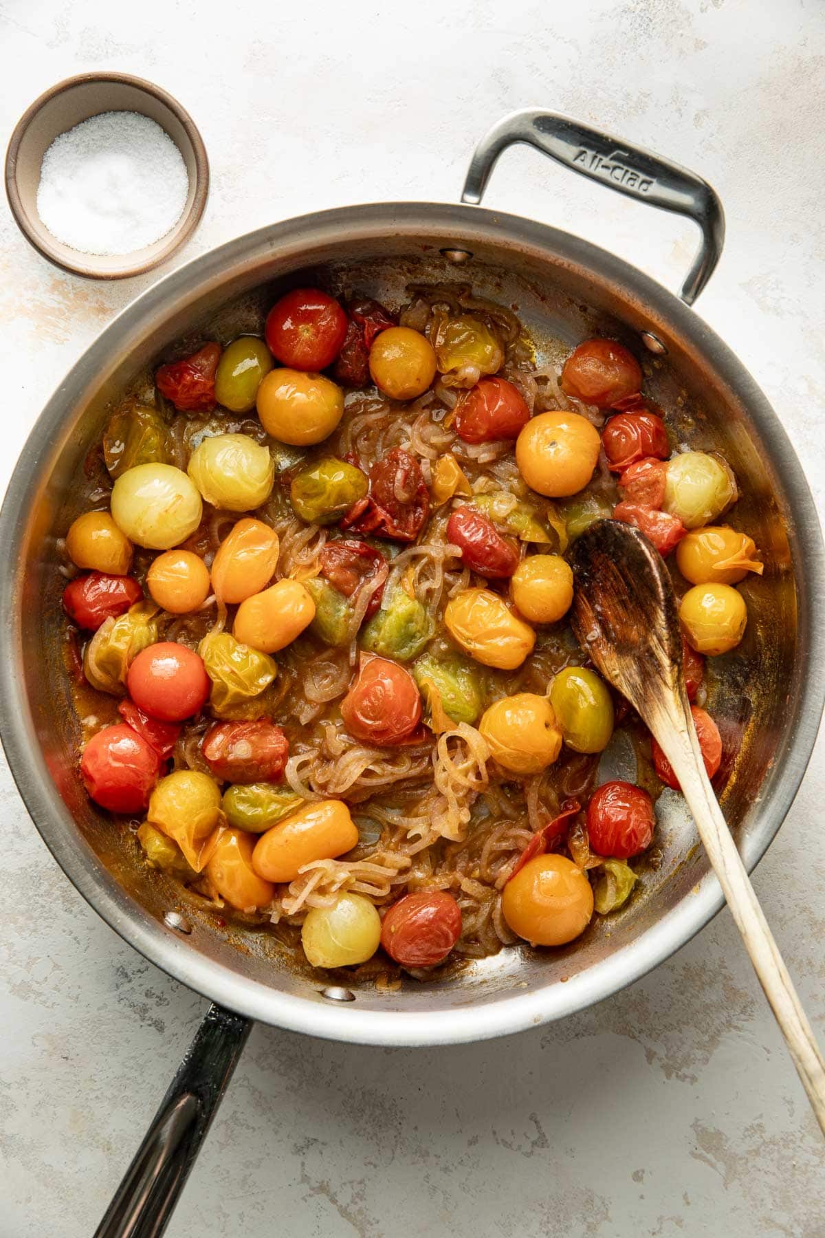 A stainless steel pan filled with sautéed cherry tomatoes in red, yellow, and orange with onions, next to a small bowl of salt and a wooden spoon resting inside the pan.