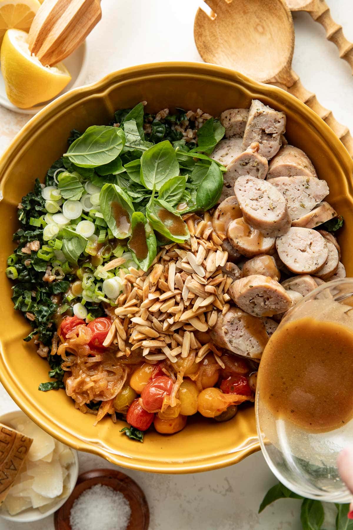 A yellow bowl filled with greens, sliced sausage, tomatoes, slivered almonds, green onions, and basil, with dressing being poured on top; wooden utensils and lemon nearby.