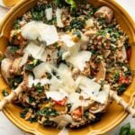 A yellow bowl filled with farro salad, kale, mushrooms, tomatoes, sausage, and topped with shaved Parmesan cheese. Two wooden serving spoons rest inside the bowl. Lemon wedges and a small bowl of salt are nearby.