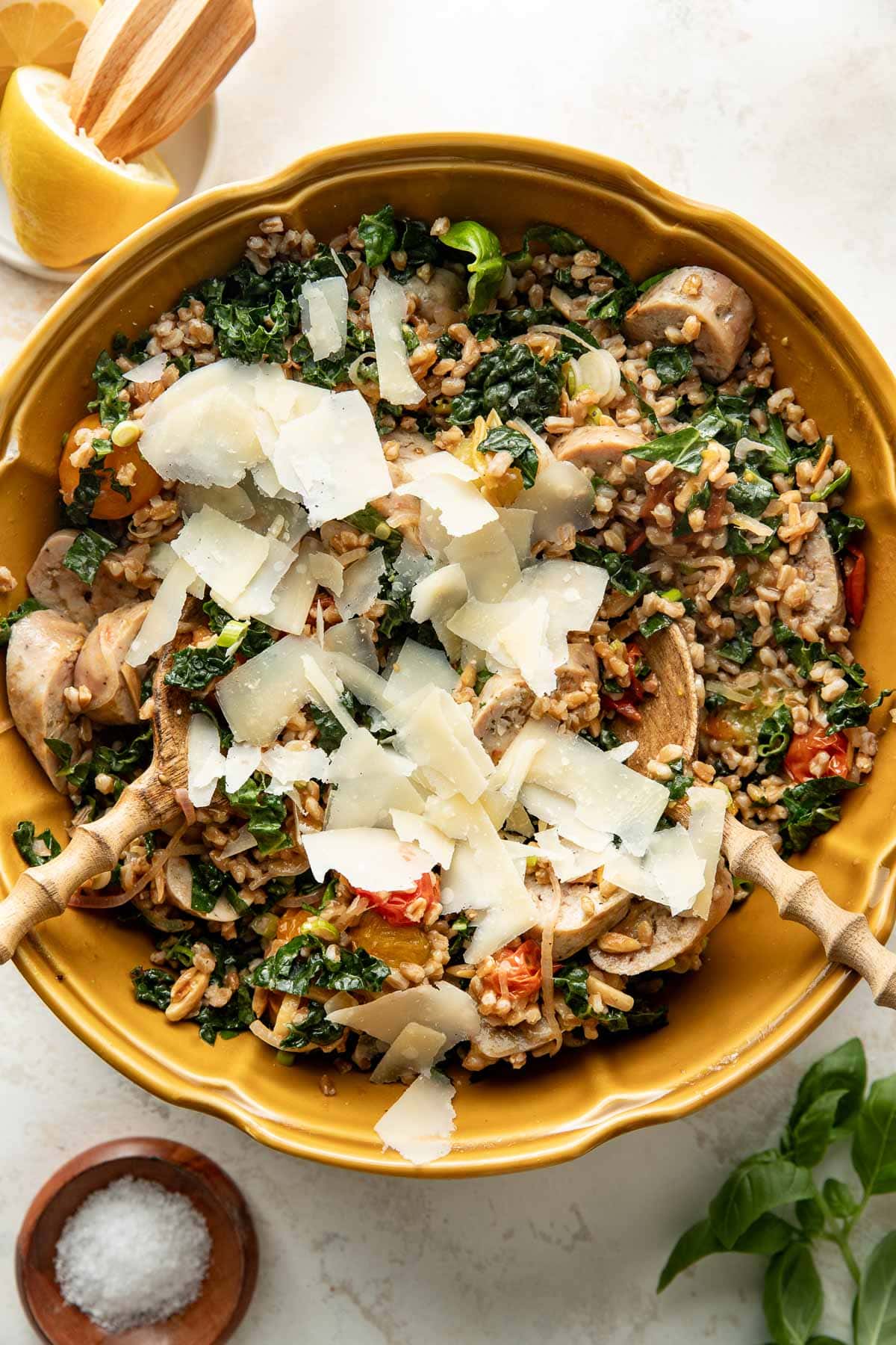 A yellow bowl filled with farro salad, kale, mushrooms, tomatoes, sausage, and topped with shaved Parmesan cheese. Two wooden serving spoons rest inside the bowl. Lemon wedges and a small bowl of salt are nearby.