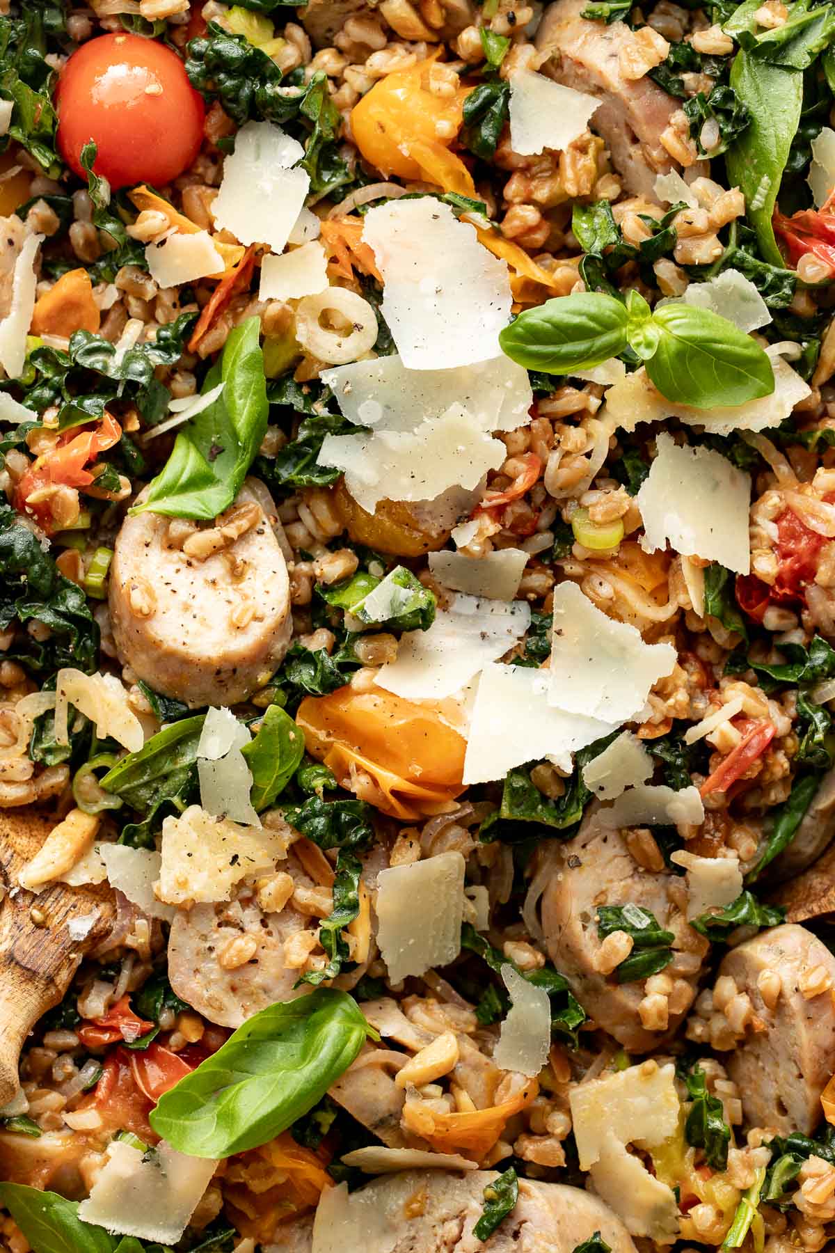 A close-up of a colorful dish featuring sliced sausage, grains, leafy greens, cherry tomatoes, fresh basil leaves, and shaved parmesan cheese.