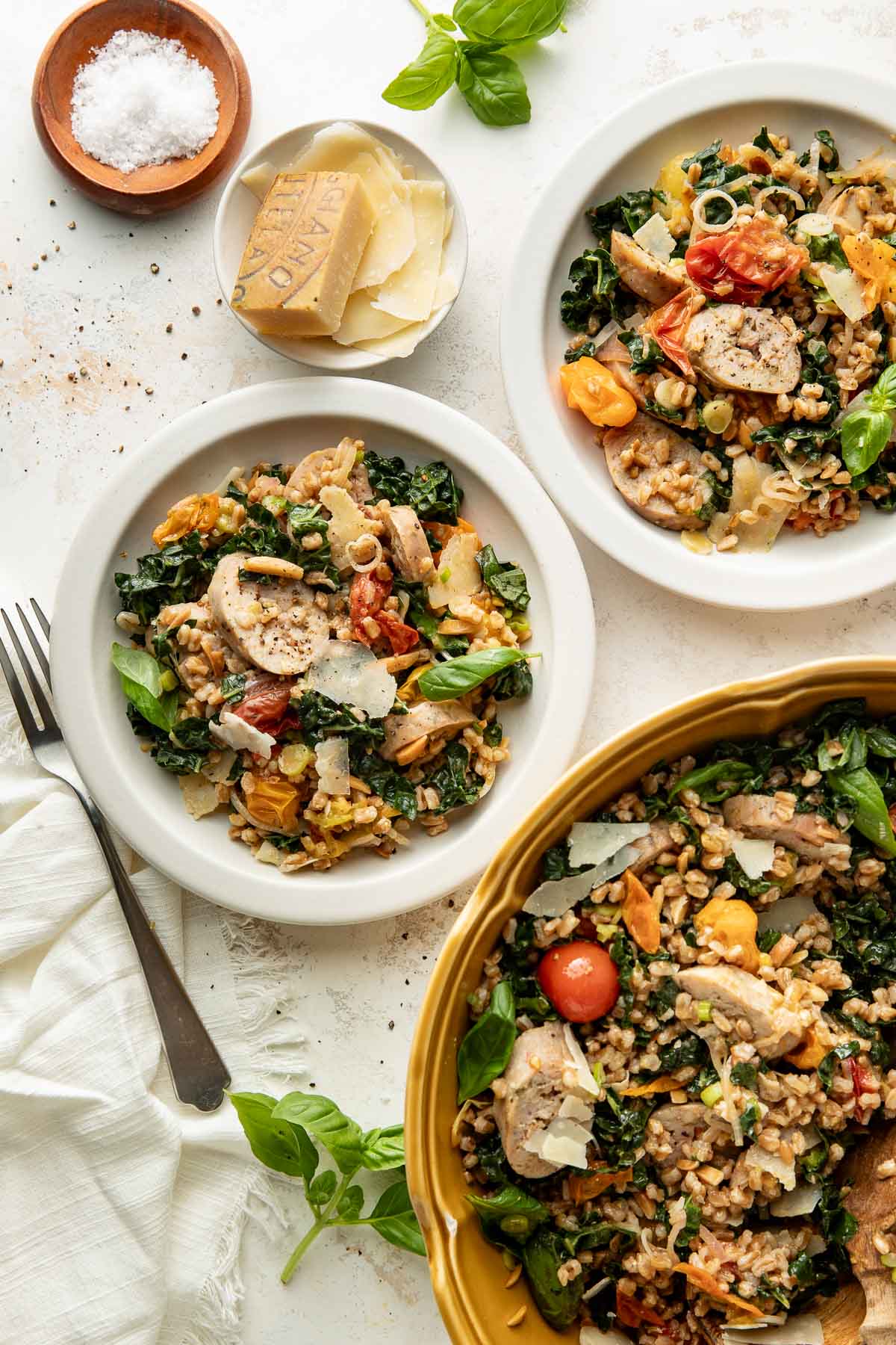 Three plates with chicken, cherry tomatoes, greens, and grains, topped with shaved cheese. A bowl of salt, a wedge of cheese, and fresh basil are nearby. The dishes are arranged on a white surface with a napkin and knife.