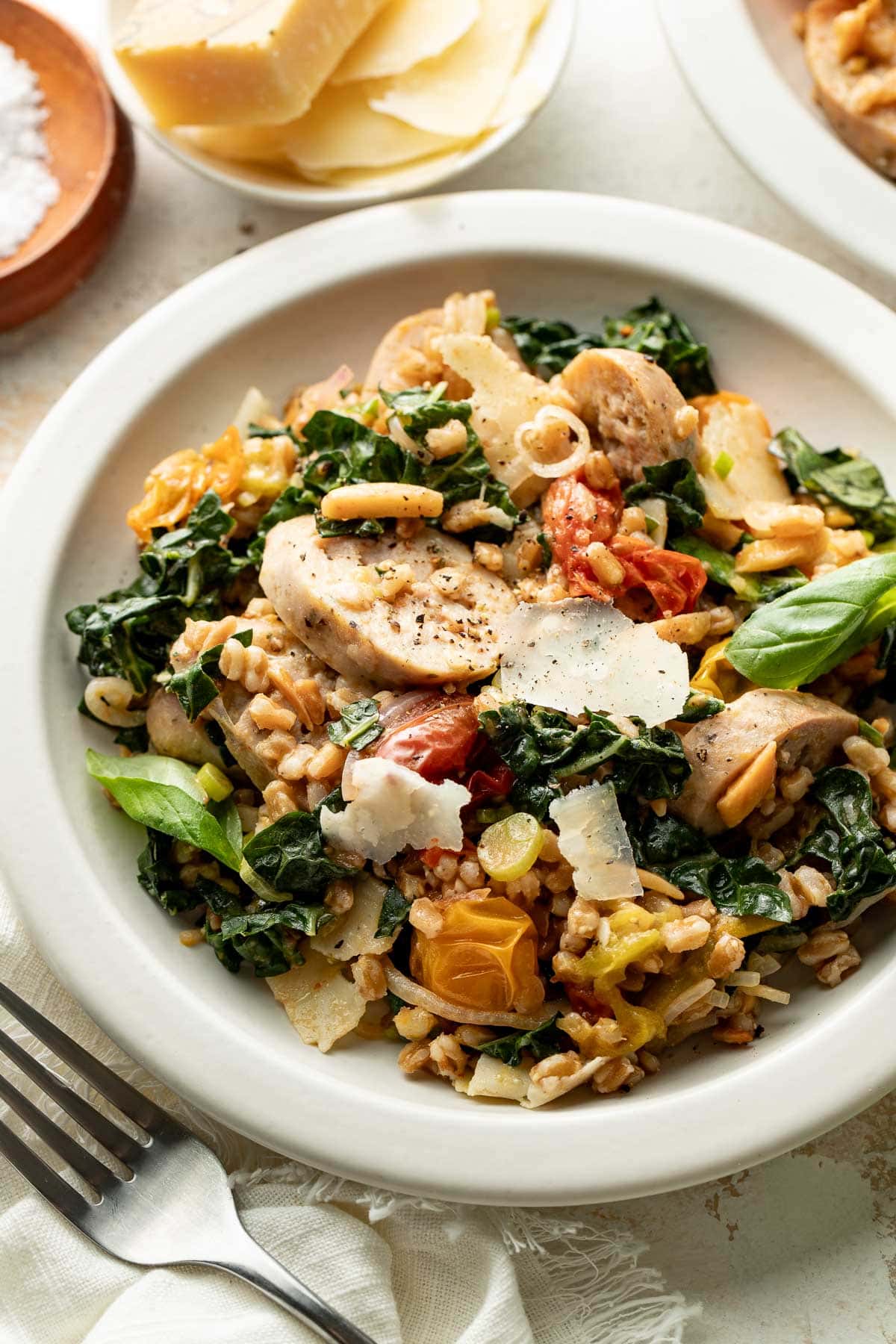 A bowl of farro salad with sliced chicken, kale, cherry tomatoes, shaved parmesan, nuts, and basil leaves, served on a white plate with a fork and cheese in the background.