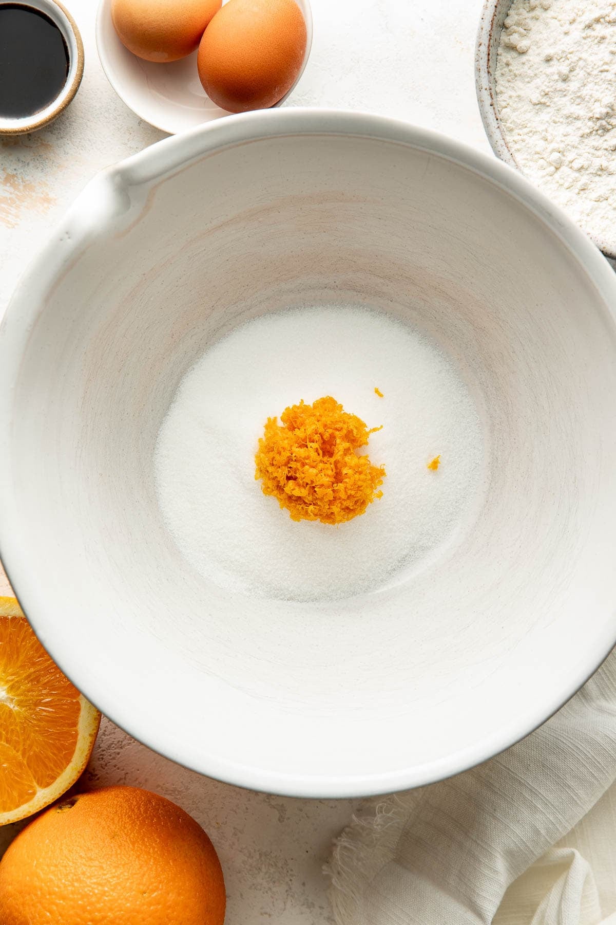 A white bowl holds sugar and orange zest. Surrounding it are eggs, flour, a small bowl of vanilla, and halved oranges, all set on a light surface.
