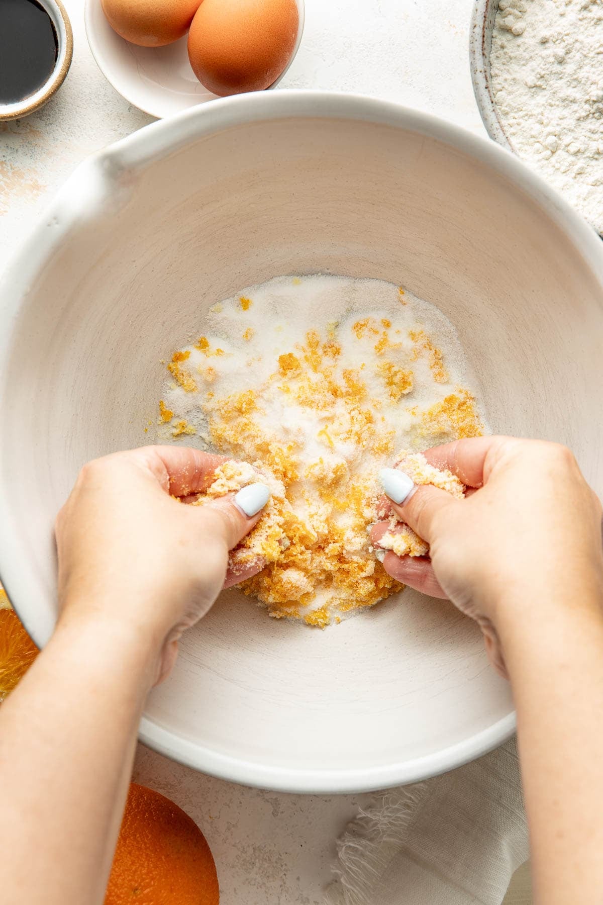 Hands rubbing orange zest and sugar together in a white mixing bowl, surrounded by baking ingredients like eggs, flour, and vanilla on a light surface.