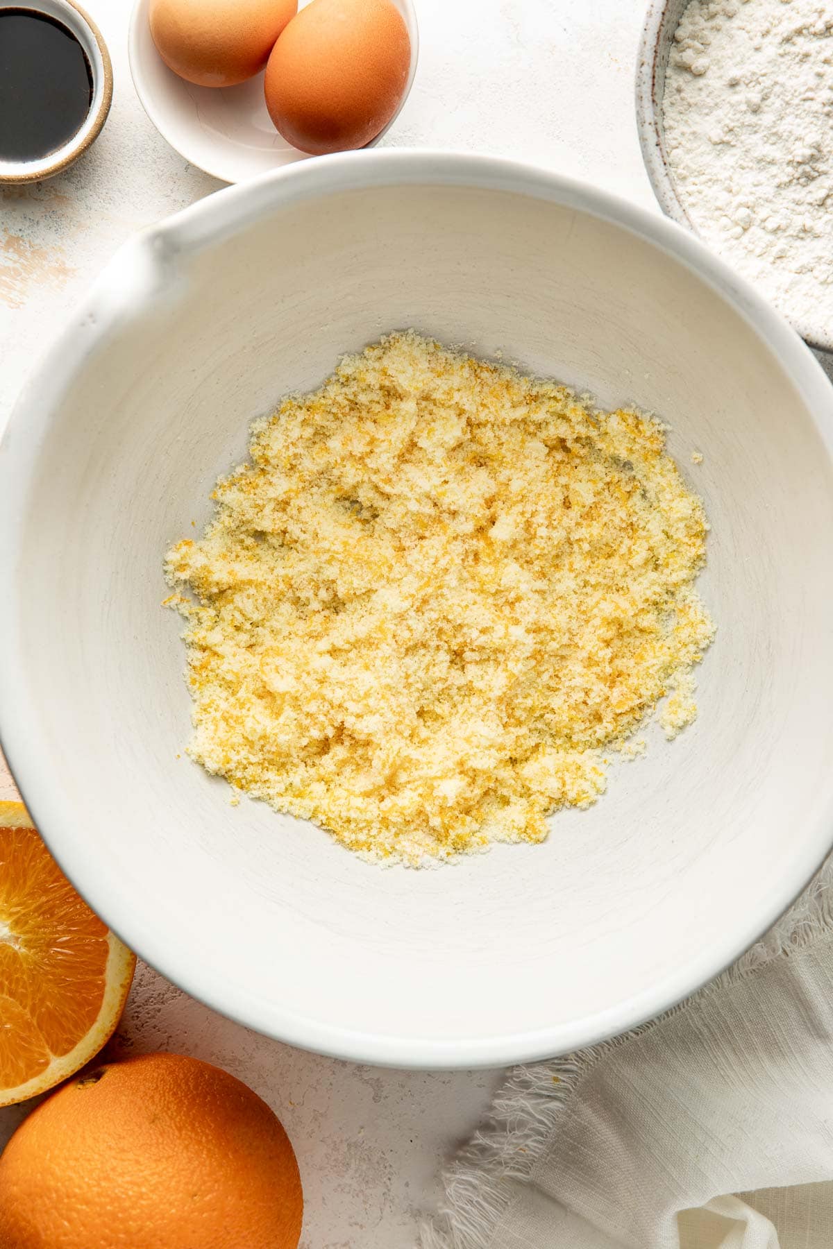 A white bowl with a yellow, crumbly mixture sits on a counter surrounded by eggs, flour, orange halves, and a small dish of dark liquid, likely vanilla extract.