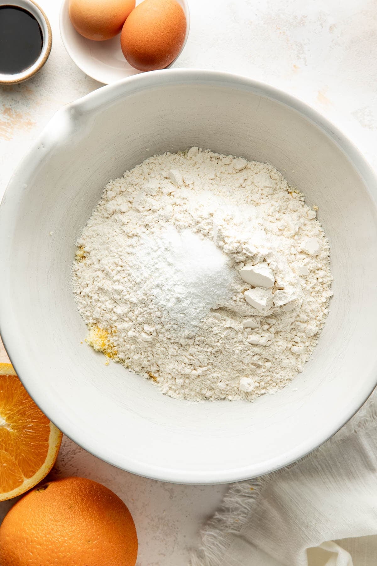 A white bowl with flour and baking soda, surrounded by eggs, an orange, vanilla extract in a small bowl, and a white napkin on a light surface.