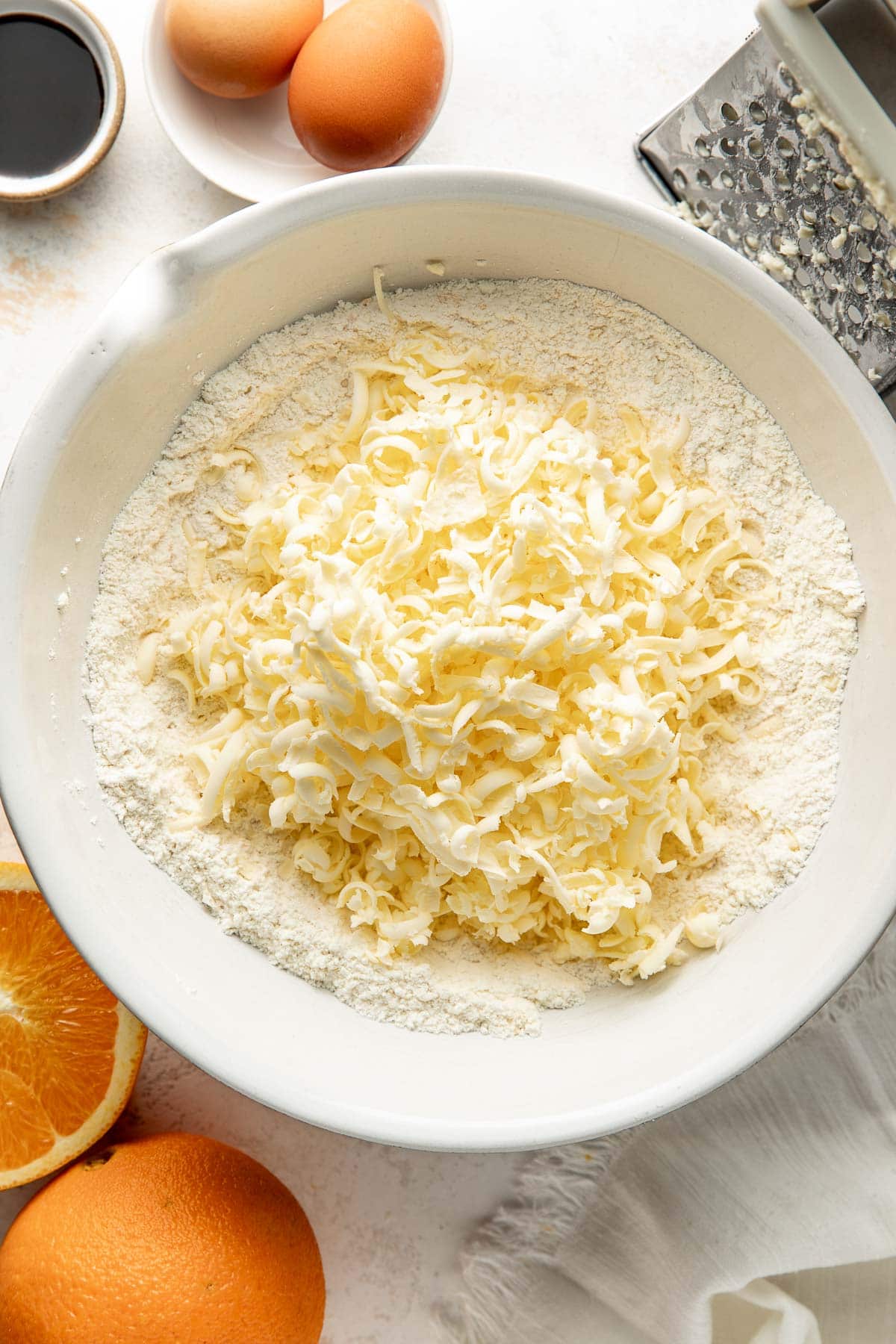 Large bowl filled with flour and shredded cheese, surrounded by whole eggs, orange halves, a cheese grater, and a small bowl of liquid on a white surface.