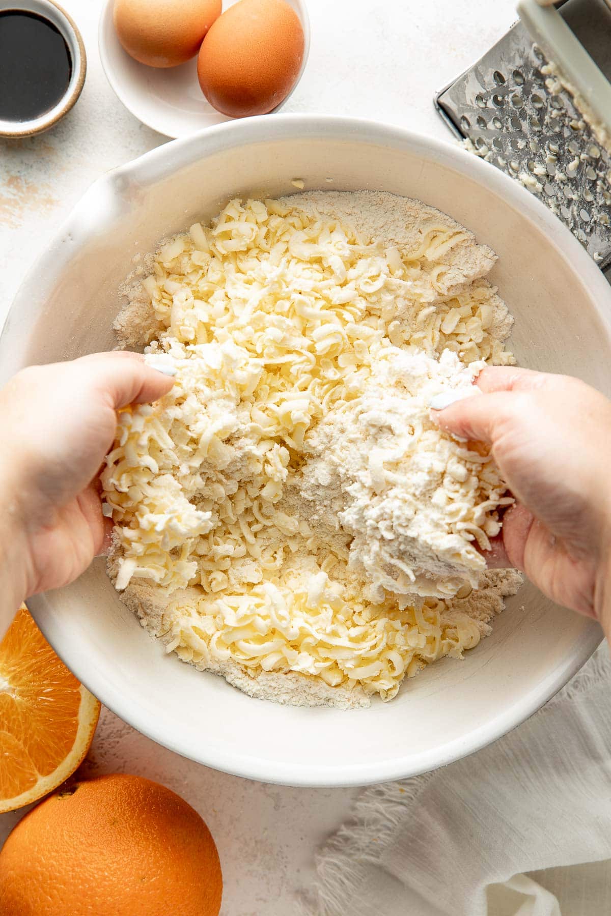 Hands mixing grated butter into flour in a white bowl. Nearby are eggs, an orange, a metal grater, and a small bowl with liquid, all set on a white countertop.