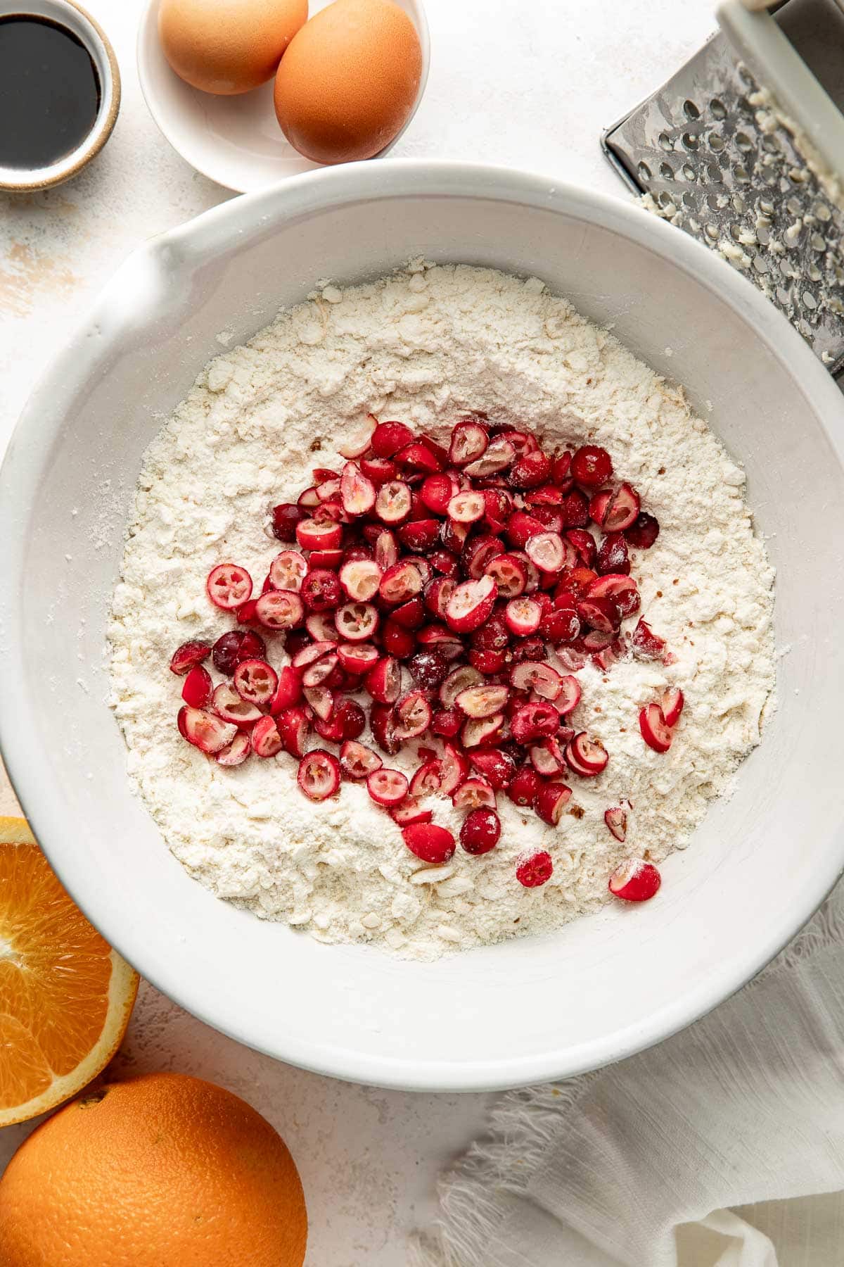 A white bowl filled with flour and chopped cranberries sits on a counter, surrounded by two eggs, an orange, a grater, and a small dish of vanilla extract.