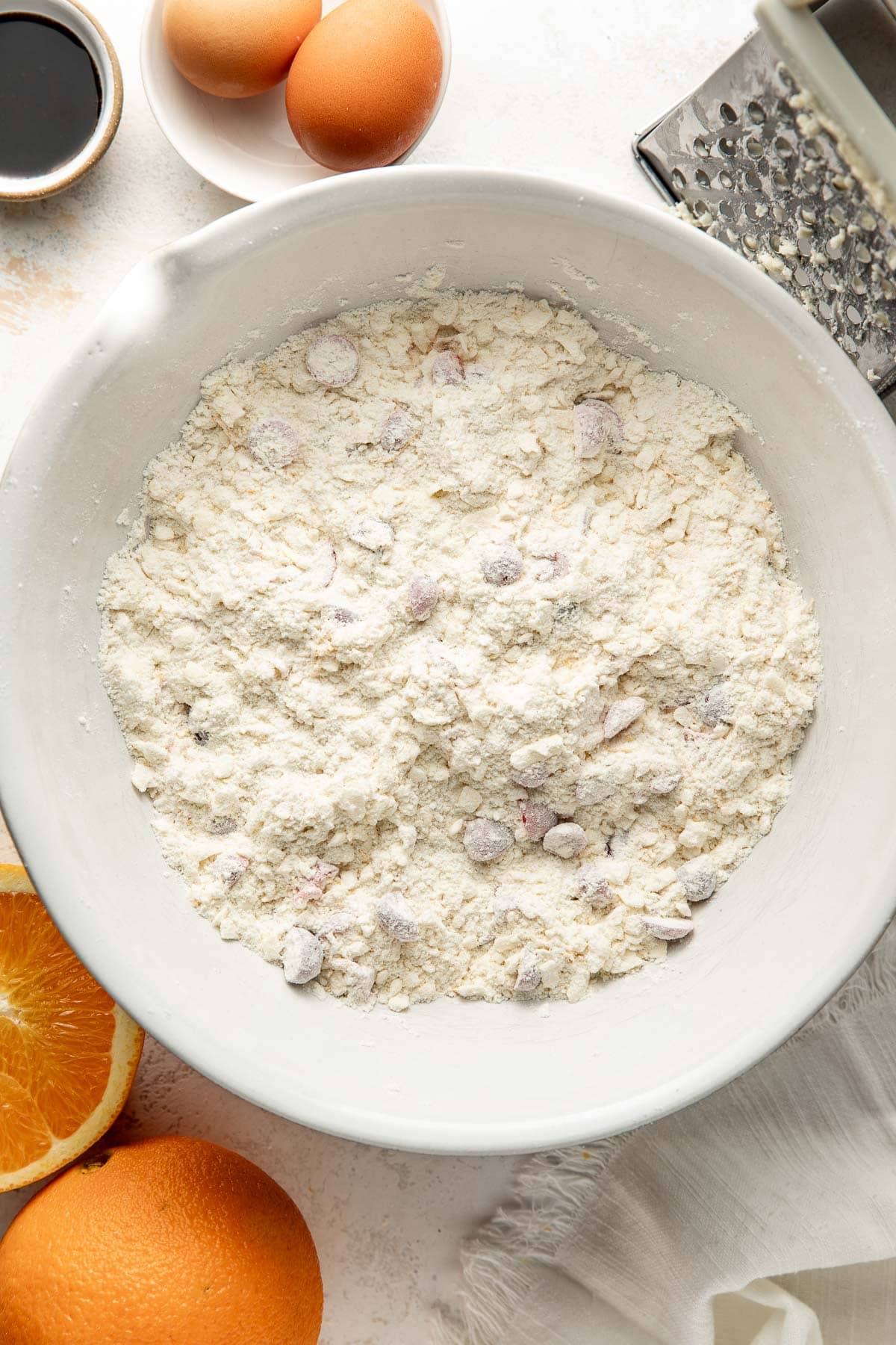 A white bowl filled with flour and chocolate chips sits on a counter near eggs, orange halves, vanilla extract, a grater, and a white napkin.