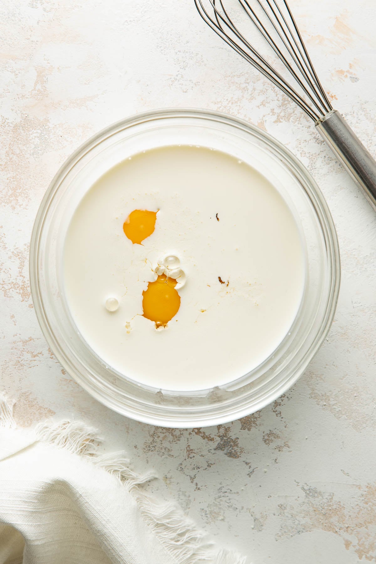 A glass bowl with cream and two egg yolks sits on a light surface near a metal whisk and a white cloth napkin.