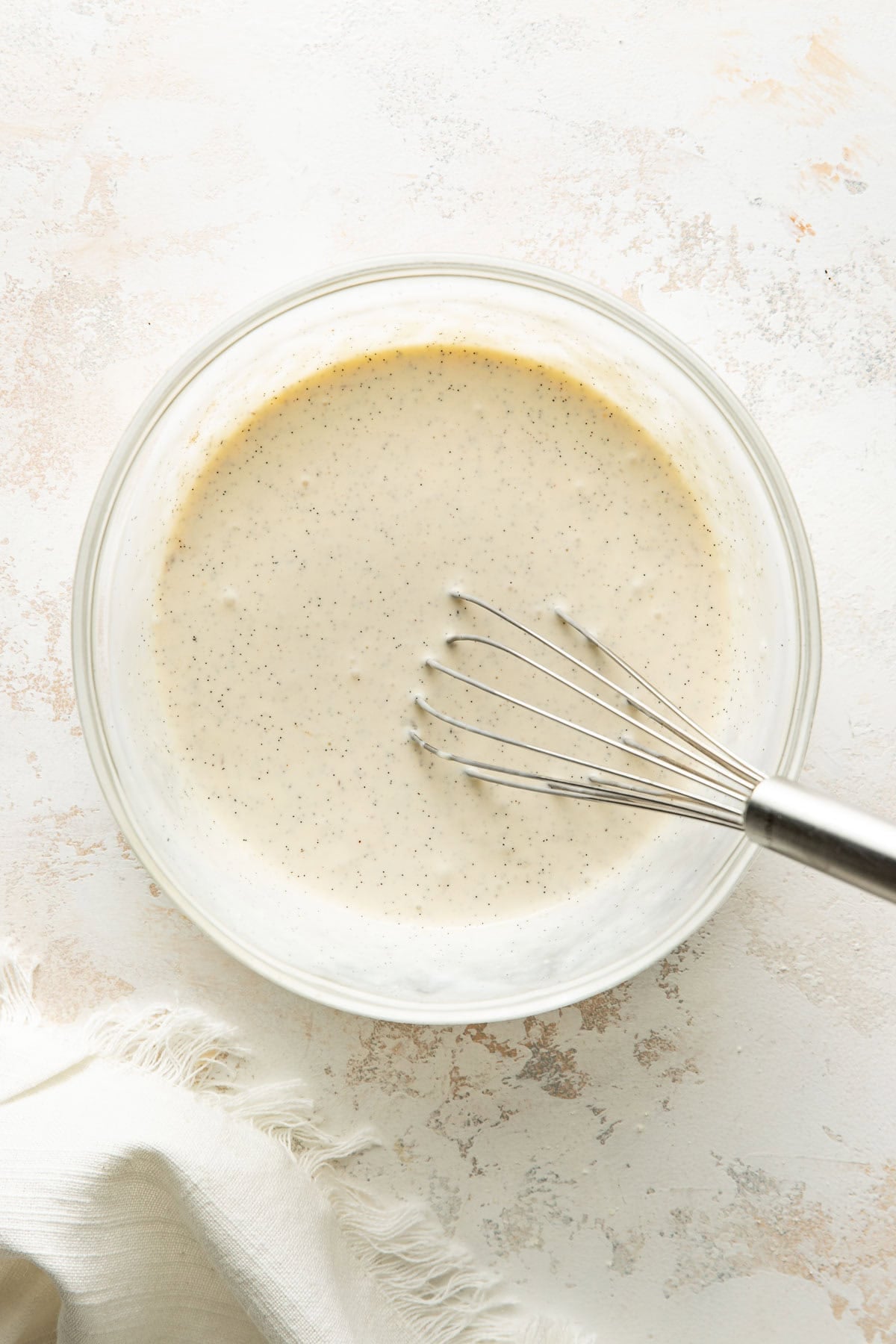 A glass bowl filled with creamy, speckled batter is being whisked with a metal whisk on a light countertop next to a white fringed cloth.
