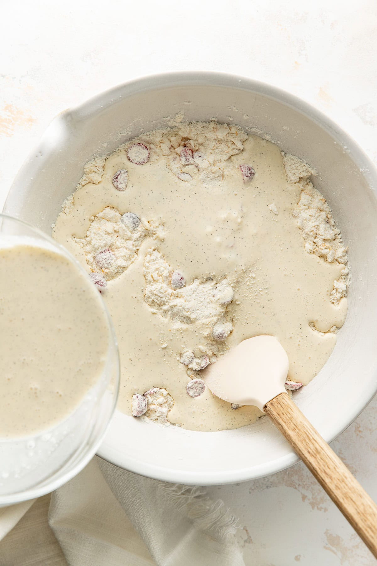A mixing bowl with flour, dried cranberries, and batter being mixed with a spatula; more batter is being poured in from a glass bowl.