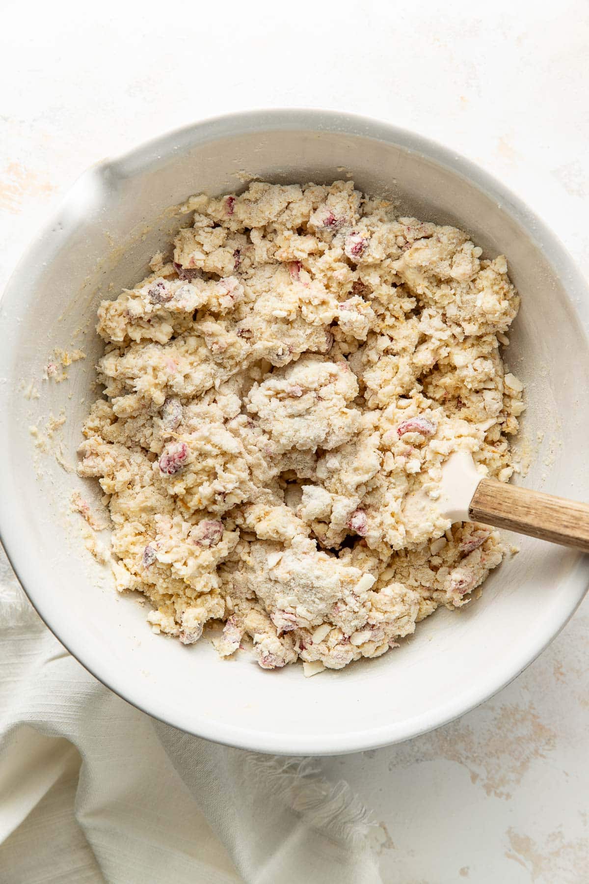 Bowl of crumbly scone dough with visible chunks and a spatula resting inside, sitting on a light surface near a cream-colored cloth.