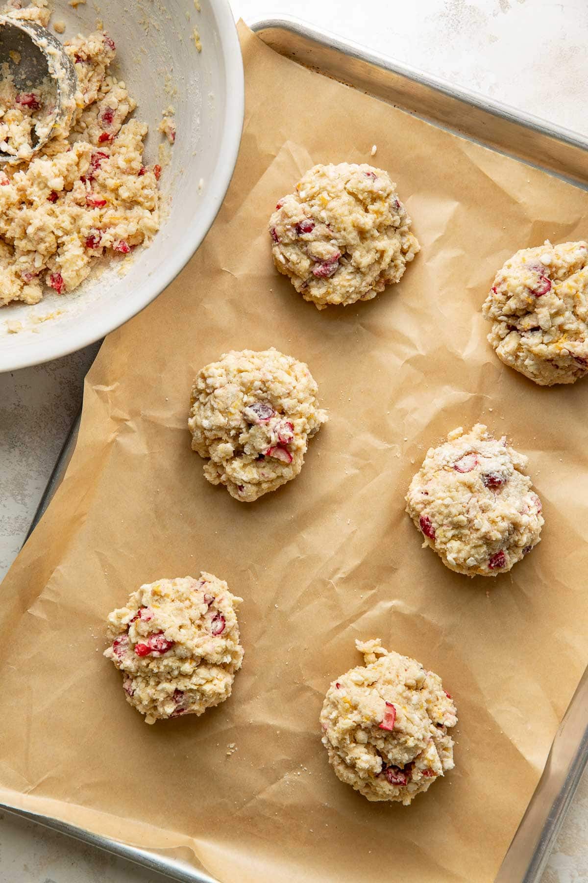 Six unbaked scones with pieces of fruit on a parchment-lined baking sheet, next to a mixing bowl with leftover dough.