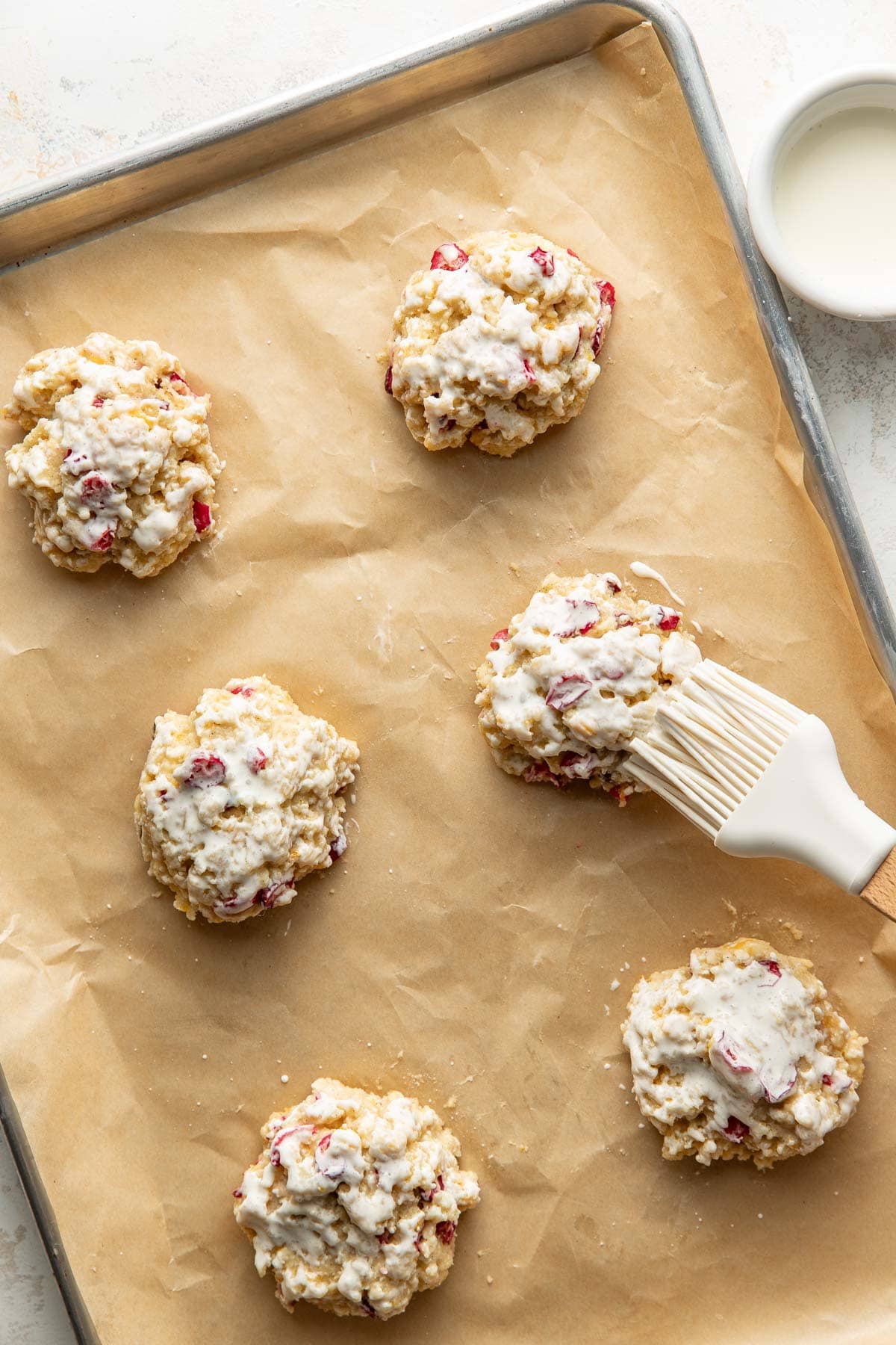 Six unbaked scones with visible red fruit pieces are on a parchment-lined baking sheet. Some have been brushed with a milky white glaze using a pastry brush. A small bowl of glaze sits nearby.