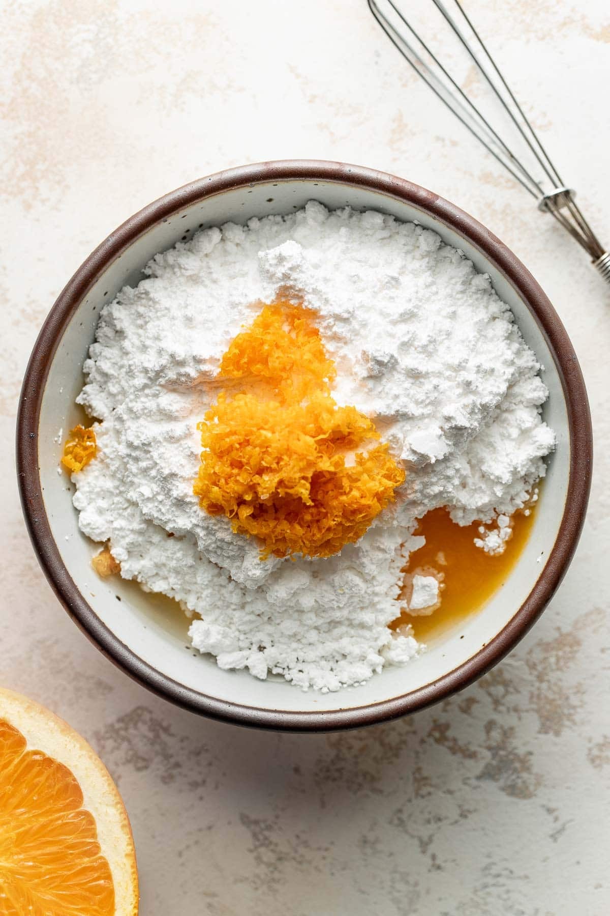 A bowl with powdered sugar, orange zest, and liquid ingredients. A whisk and a halved orange are nearby on a light surface.