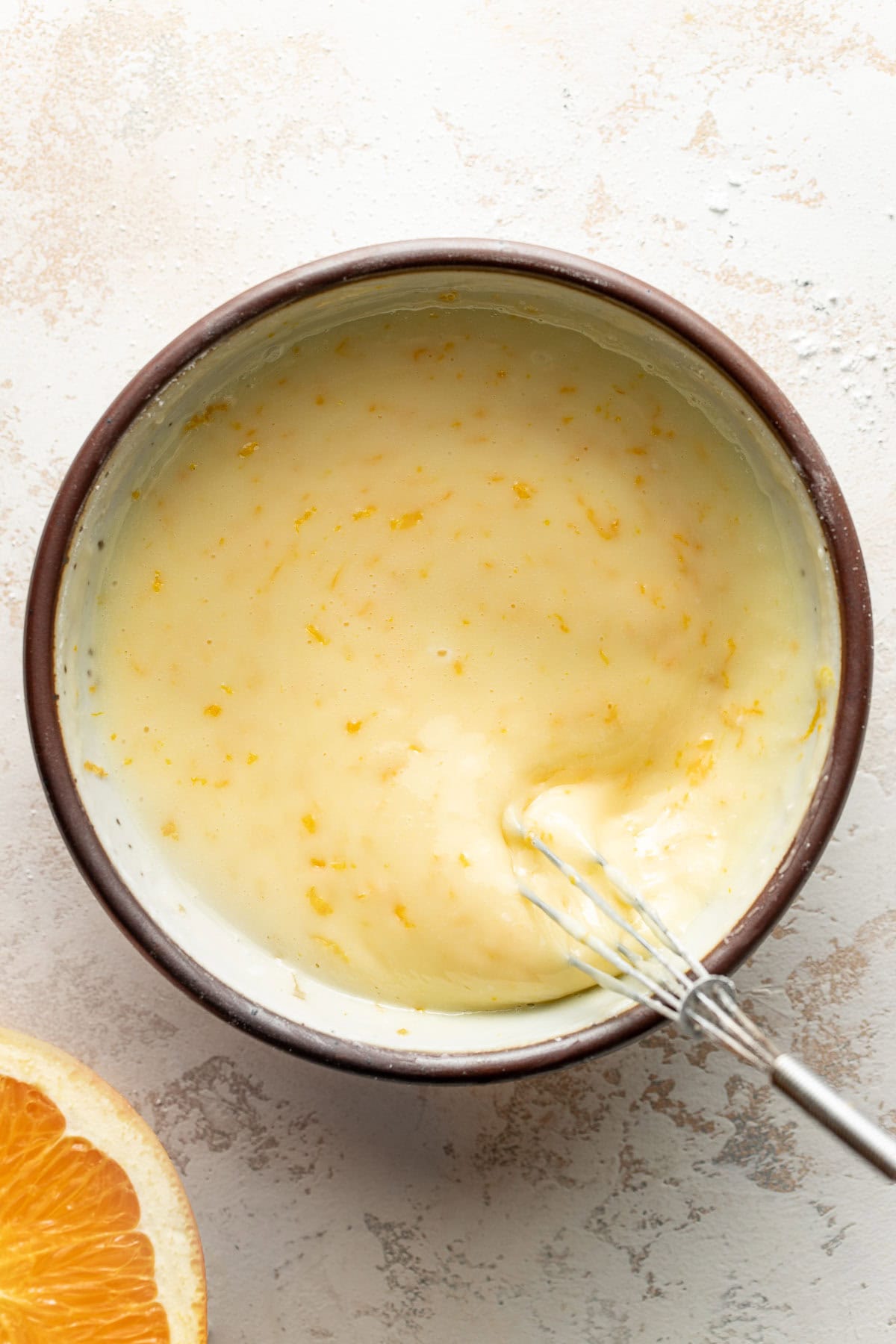A bowl of creamy mixture with orange zest, being whisked. A metal whisk rests inside the bowl, and a partially visible orange sits nearby on a light, textured surface.