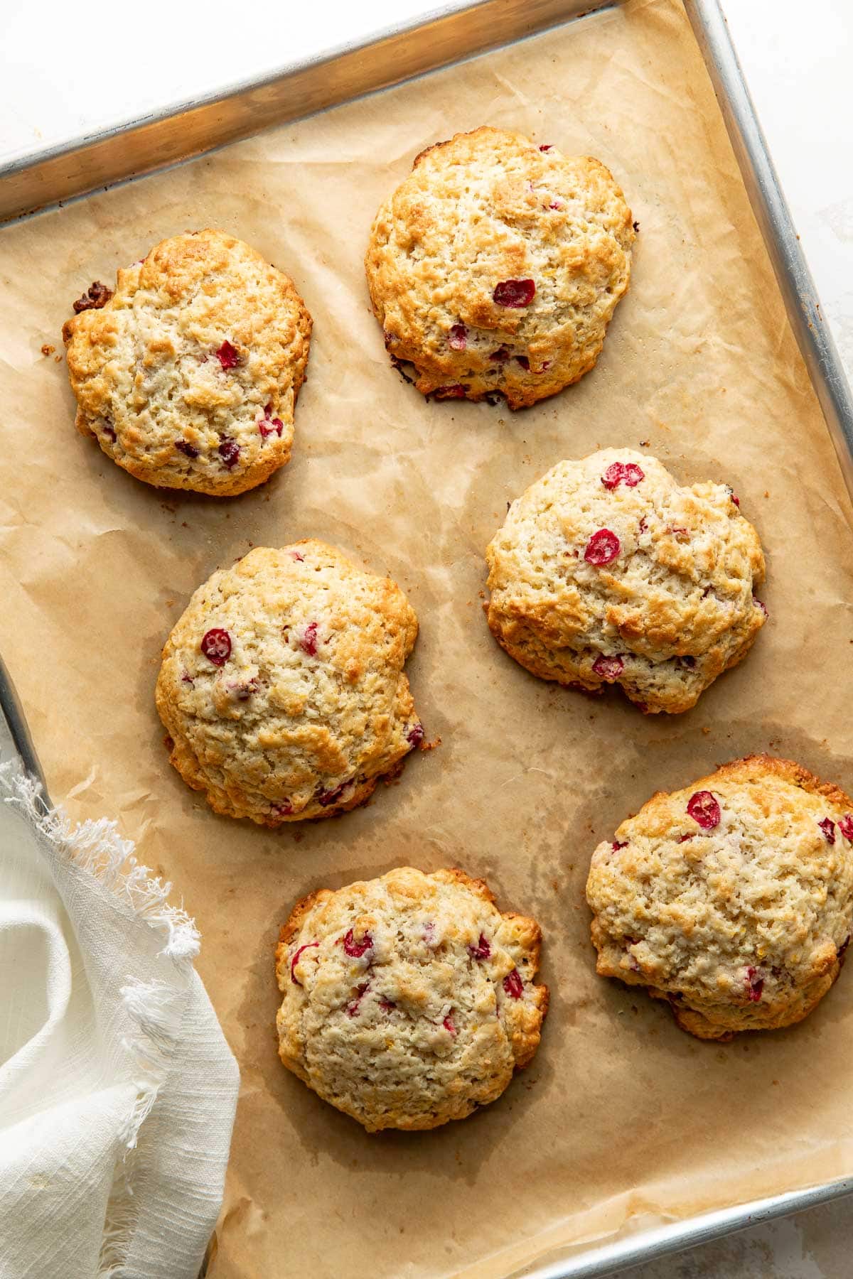 Six golden brown scones with visible cranberries rest on a parchment-lined baking sheet, viewed from above. A white cloth napkin is partially visible in the corner.