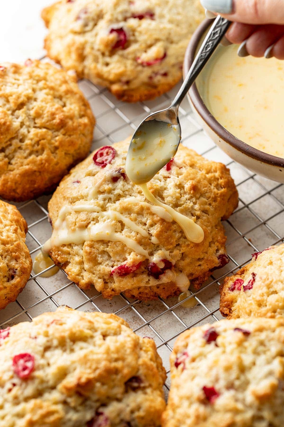 A hand drizzles glaze over freshly baked cranberry scones on a wire rack, with a bowl of glaze beside them.