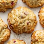 Close-up of golden-brown scones with visible cranberries, drizzled with a light white glaze, arranged closely together on a textured, off-white surface.