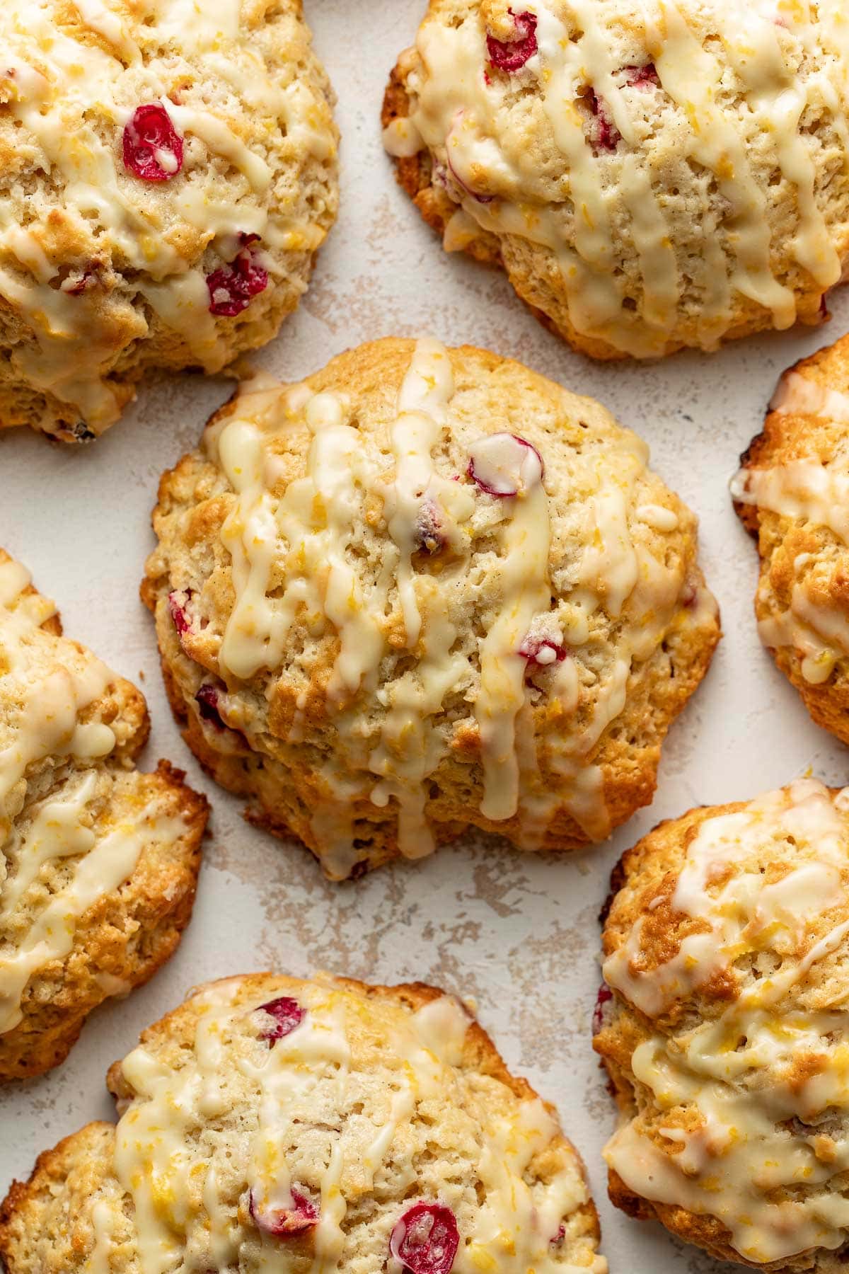 Close-up of golden-brown scones with visible cranberries, drizzled with a light white glaze, arranged closely together on a textured, off-white surface.