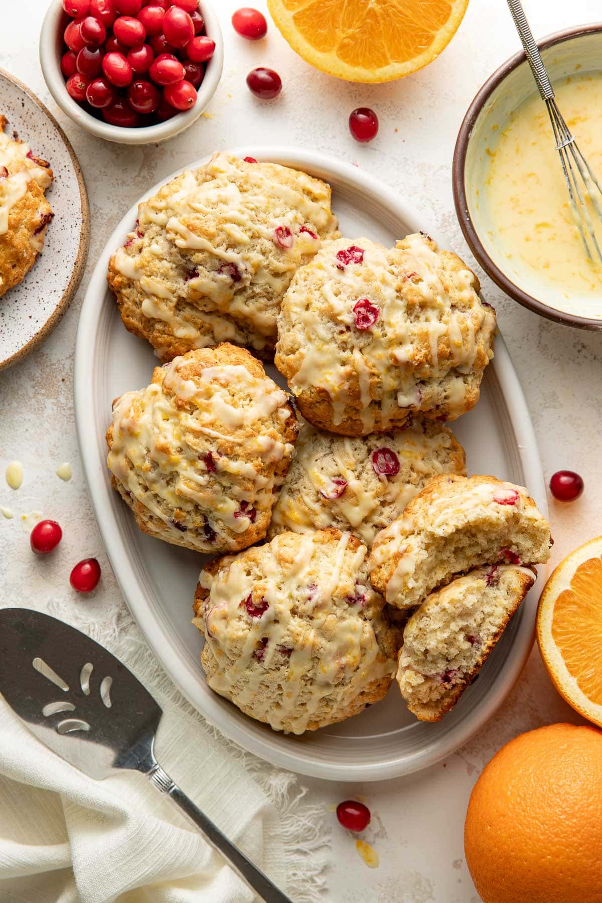 A plate of cranberry orange cookies drizzled with icing, surrounded by fresh cranberries, orange halves, a bowl of icing, a spatula, and a napkin on a light background.