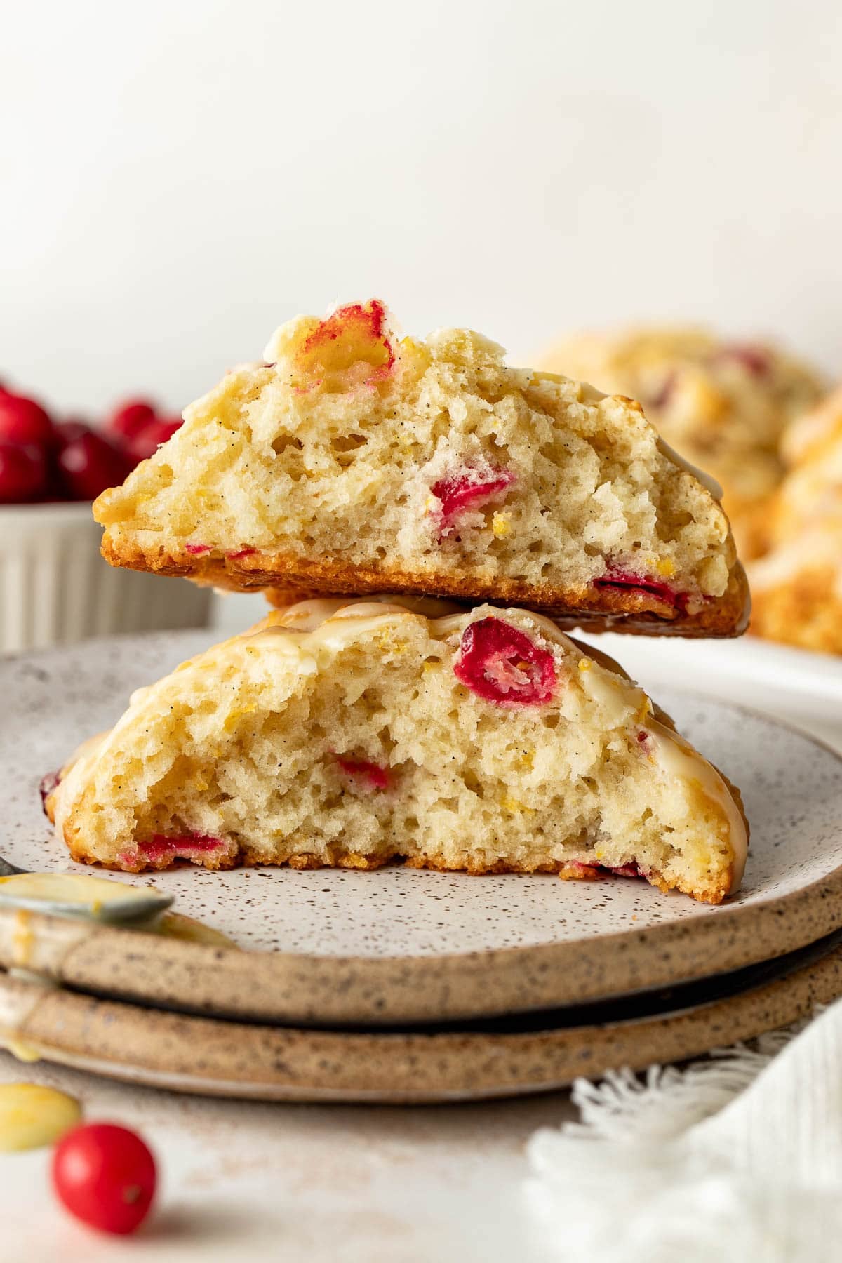 Two halves of a cranberry scone are stacked on a plate, showing a moist, crumbly interior with visible red cranberries. A bowl of cranberries and more scones are blurred in the background.