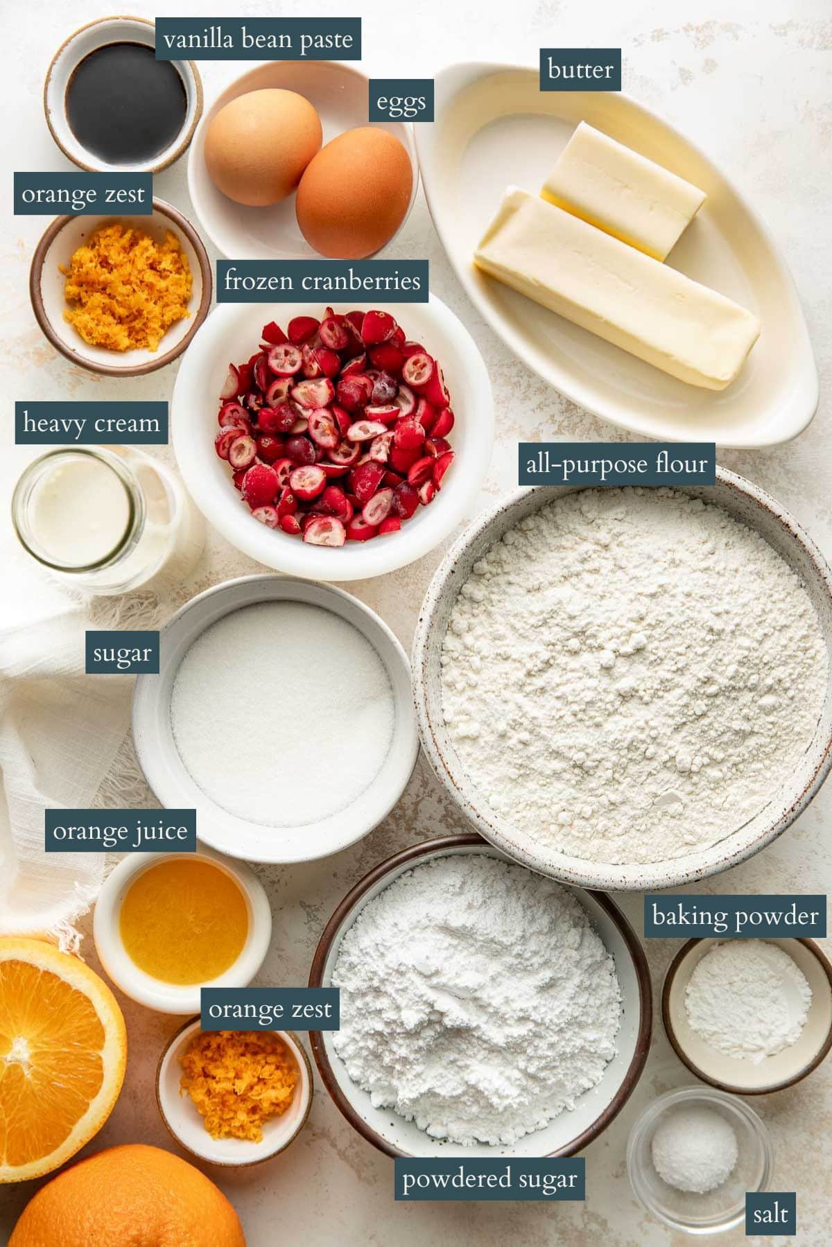 Bowls and plates with labeled baking ingredients: butter, eggs, vanilla bean paste, orange zest, frozen cranberries, heavy cream, all-purpose flour, sugar, orange juice, powdered sugar, baking powder, and salt.