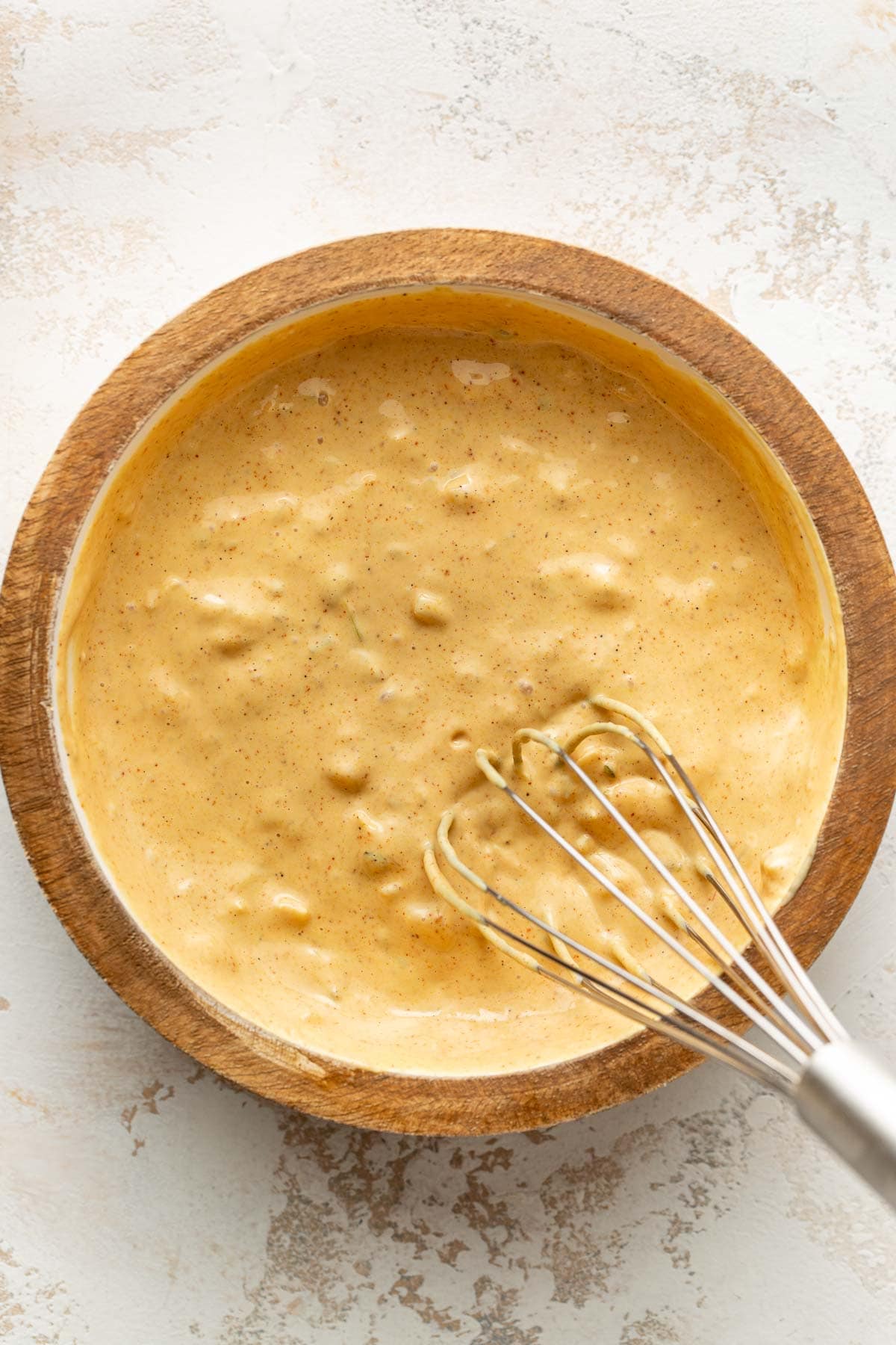 A wooden bowl filled with a thick, creamy yellow batter, speckled with spices, sits on a light surface. A metal whisk rests inside the bowl, partially coated with the batter.